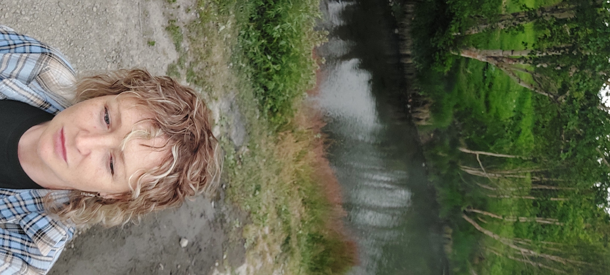 The image shows a woman with blonde, curly hair and freckles, looking down and to the side. She is wearing a blue and white plaid shirt over a black t-shirt and a small hoop earring in her left ear. The background features a natural landscape with trees, a body of water, and green foliage. The location is identified as Nachiki, Russia. The scene appears to be outdoors, possibly near a river or stream, with a sense of natural beauty and tranquility. The lighting suggests it is daytime, possibly overcast.