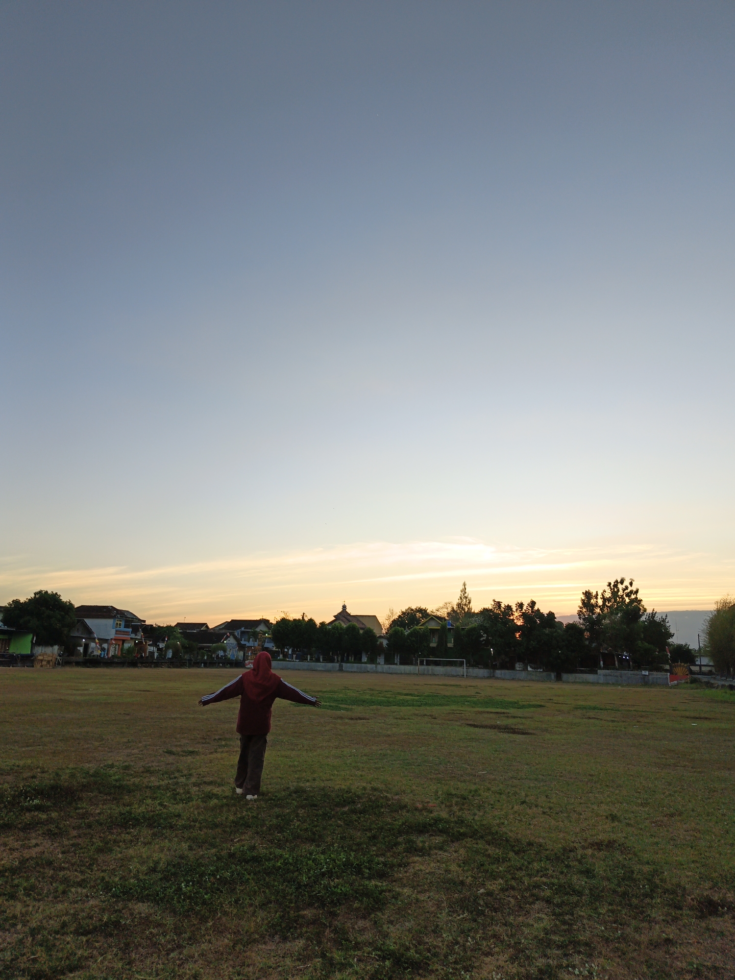 A person wearing a red hooded jacket and brown pants stands in a grassy field with arms outstretched. The person is facing away from the camera, towards a line of buildings and trees in the distance. The sky above is a gradient of light blue to pale orange, indicating either sunrise or sunset. The field appears to be a sports field, with what looks like a goalpost visible on the left side of the frame. The scene suggests a tranquil moment in an open area, possibly a park or school grounds in Klaten, Indonesia, during the early morning or late evening.
