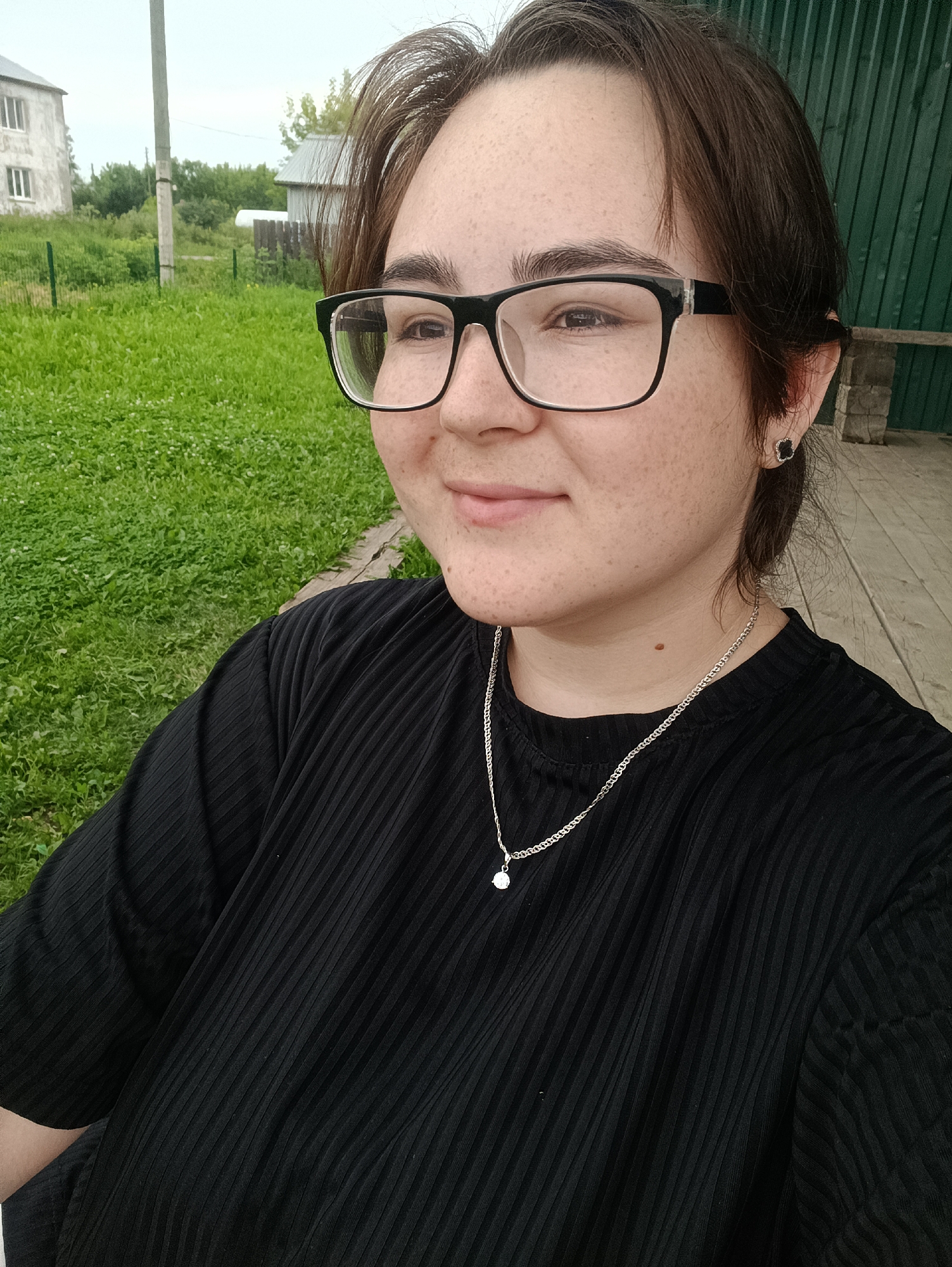 A young person wearing black-rimmed glasses, a black ribbed t-shirt, and a silver necklace with a pendant is pictured. They have brown hair and a fair complexion with freckles. They are wearing a dark stud earring in their left ear. The background suggests an outdoor setting in a rural or suburban area, possibly in or near the city of Posad, Russia. To the left, there is a grassy field with a white building in the distance and a utility pole. To the right, a green wooden structure resembling a fence or building wall is visible, with a wooden bench or platform. The ground is covered with grass and some patchy dirt. The lighting suggests an overcast day.