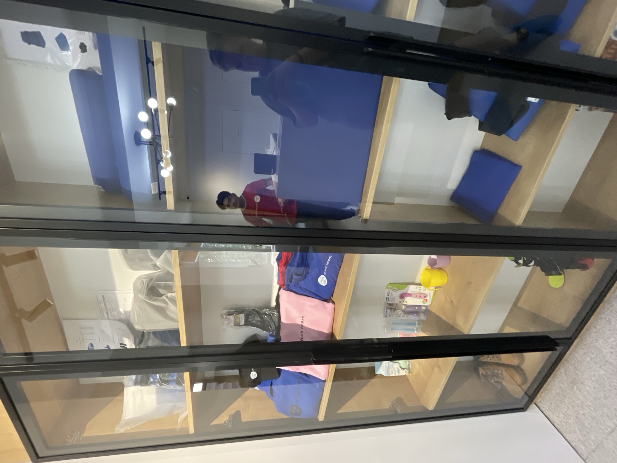 The image shows a display cabinet with glass doors, likely in a retail or service establishment. The cabinet is filled with various items, including blue and pink bags with "rehab team" printed on them, a black cap, socks in packaging, and what appear to be toiletries such as toothbrushes and small cups on a lower shelf. Two pairs of boots are visible on a shelf in the bottom left section of the cabinet.
Through the glass, two individuals are partially visible. One, wearing a red t-shirt with yellow stripes, stands behind a blue counter. Another person, reflected in the glass, appears to be taking a photograph with a smartphone. The background suggests an indoor setting with lighting fixtures visible on the ceiling of the upper section of the cabinet.
The scene appears to be in Almaty, Kazakhstan, given the provided metadata, though no direct visual cues within the image confirm this location. The overall impression is a well-organized display of merchandise or promotional items.