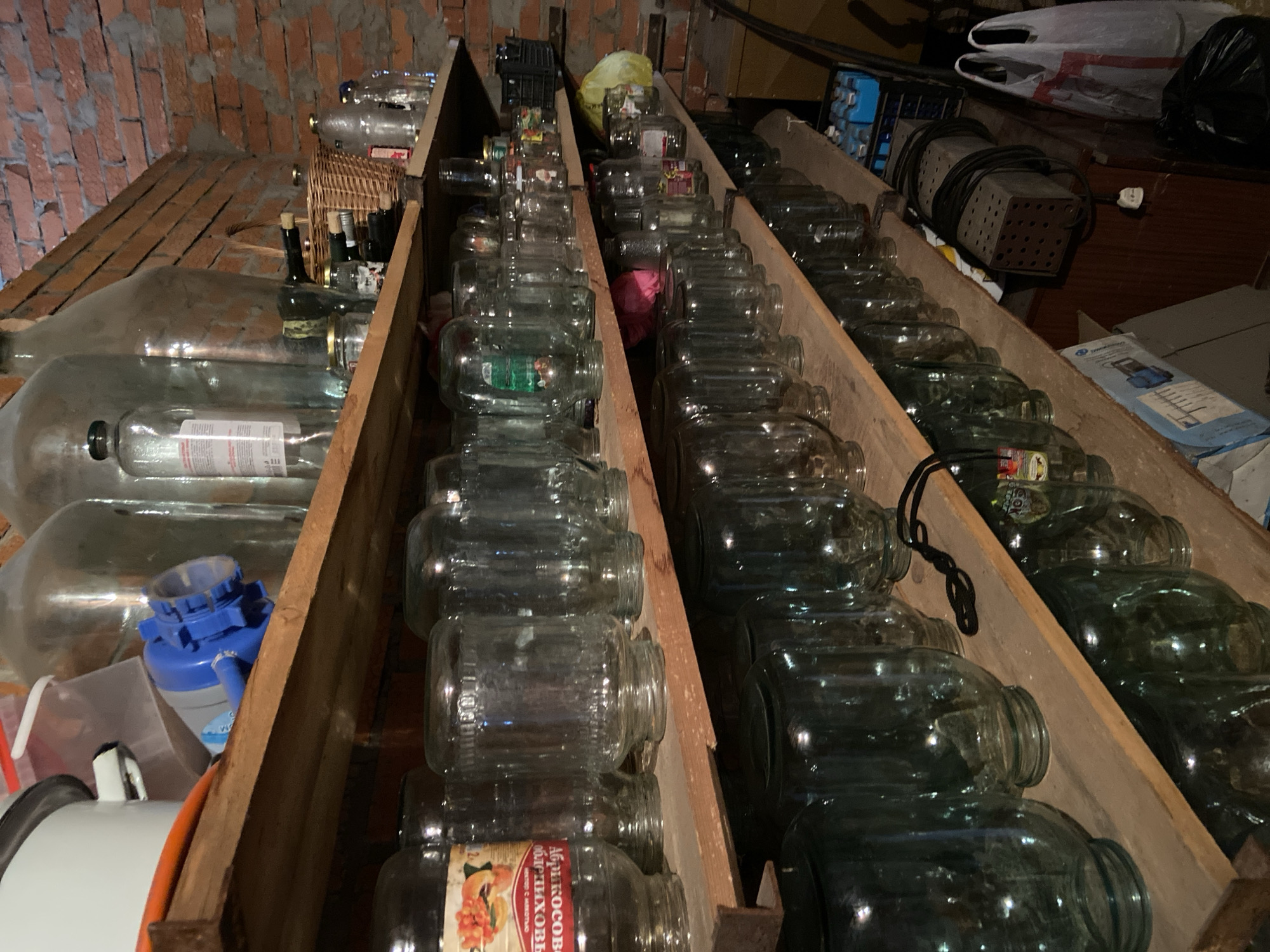 The image depicts an interior storage space, possibly a cellar or pantry, with three rows of wooden shelving. The shelves are filled with glass jars, mostly empty, arranged in rows. Some of the jars have labels or remnants of labels in Cyrillic script. Other objects, including bottles, a basket, and various unidentified items, are also visible on the shelves. The background shows a brick wall and additional stored items. There is no evidence indicating this location is in Chaikovsky, Russia.