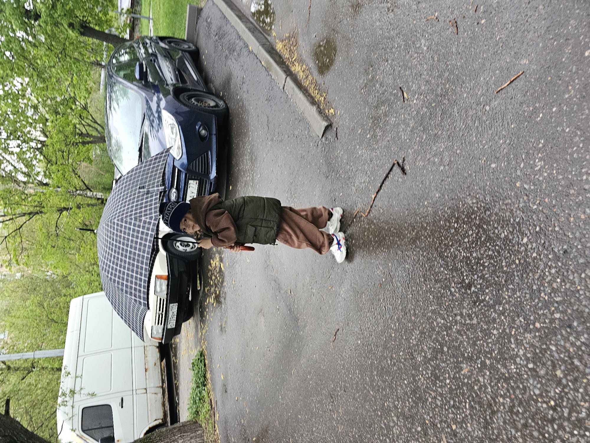 A child stands under a plaid umbrella on a wet paved road in Khimki, Russia. The child is wearing a brown tracksuit, an olive green vest, and white sneakers with blue stripes. Behind the child are parked vehicles: a blue car and a white van. Tree foliage is visible in the background. There are small sticks on the wet pavement.