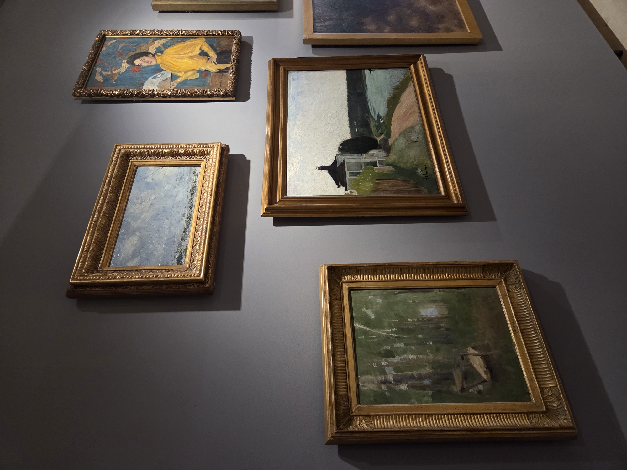 The image shows an art exhibition in Malmö, Sweden, likely at Moderna Museet Malmö, given the contemporary art and minimalist architectural style. The exhibition features a collection of framed paintings displayed on a dark gray wall.
In the foreground, on the left, a square-format painting depicts a forest scene with tall, slender trees and dappled light filtering through the leaves. A wooden structure, possibly a bench or part of a makeshift shelter, is visible within the trees. The painting is enclosed in an ornate, golden frame.
To the right and slightly above this, a horizontal painting shows a wide, pale blue sky over a body of water, with a suggestion of a shoreline in the distance. This artwork is also in a decorative golden frame.
Further to the right and higher up, a vertical painting features a woman in a bright yellow garment holding a fan. The background is a vibrant blue with stylized figures of birds and floral elements. This painting has a detailed golden frame.
Below and to the right of the forest scene, a larger horizontal painting displays a house with a dark roof and white walls, situated on a hill overlooking a body of water. A dirt path leads towards the house, and trees line the path and the water's edge. The signature "HILL" is visible in the lower right corner. This painting is in a simpler, wooden frame.
To the far right, a portion of another painting is visible, appearing to be a darker, possibly abstract or landscape composition with a muted color palette, in a simple wooden frame.
The lighting in the exhibition space is dim, with spotlights from above highlighting the artworks, creating shadows on the wall behind them. The overall atmosphere is quiet and contemplative, typical of an art gallery setting.