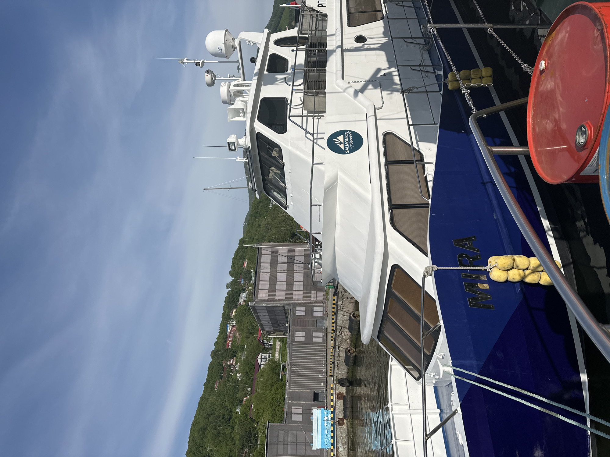 The image shows a white and blue boat docked at a pier in Petropavlovsk-Kamchatsky, Russia. The boat is named "MIRA" and has a "SALMONICA Travel" logo on its side. In the background, there are industrial buildings and a green, tree-covered hillside with houses. The sky is blue with some wispy clouds. The water is calm, reflecting the sky and the boat. There are several yellow buoys attached to the side of the boat and a red barrel is visible in the foreground. The scene appears to be a sunny day in a harbor or port area.