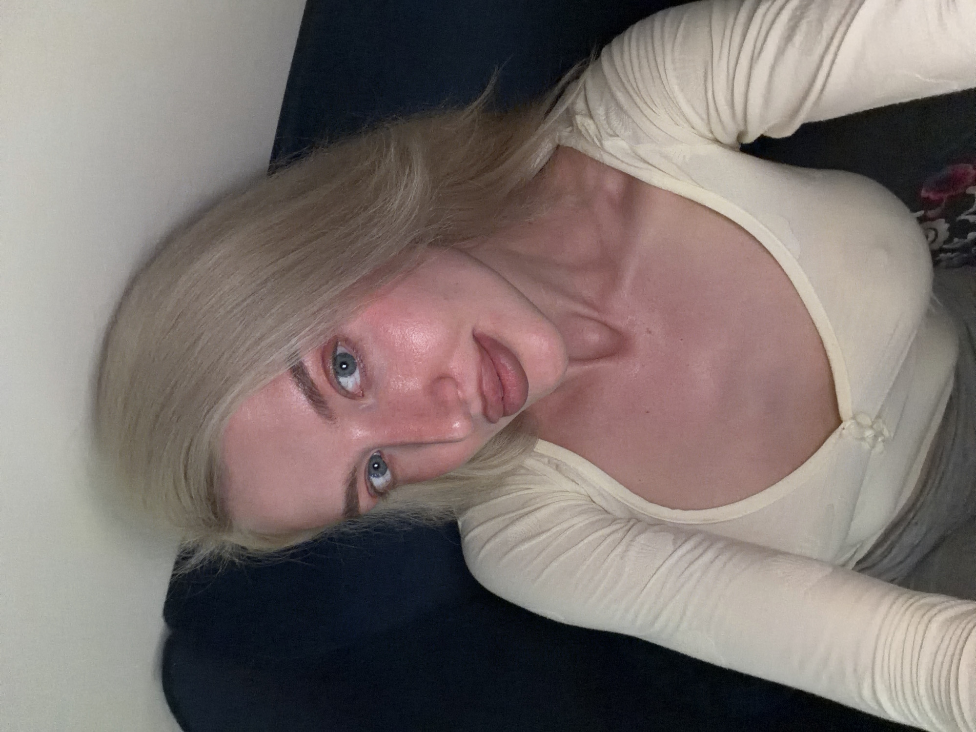 A young woman with blonde hair and blue eyes is captured in a close-up, angled shot, likely a selfie, indoors. She has fair skin, with a noticeable flush on her face and neck. Her blonde hair frames her face, and she is looking directly at the camera with a relaxed expression, featuring a faint smile or smirk.
She is wearing a long-sleeved top, light-colored (appearing cream or light yellow), with a v-neckline. Two small, matching bows or buttons are visible decorating the front of her top near the bust. Part of her left arm, covered by the textured sleeve, is visible in the foreground. A glimpse of grey fabric, possibly from pants or a skirt, can be seen to her right. She appears to be reclining or lying on a dark blue surface, which could be a couch or bed. The background on the right side includes a dark patterned fabric, possibly with a floral design, while the left side shows a plain light-colored wall. The lighting appears artificial and soft. There are no specific visual cues within the image to indicate the location as Belgrade, Serbia, and no visible text.