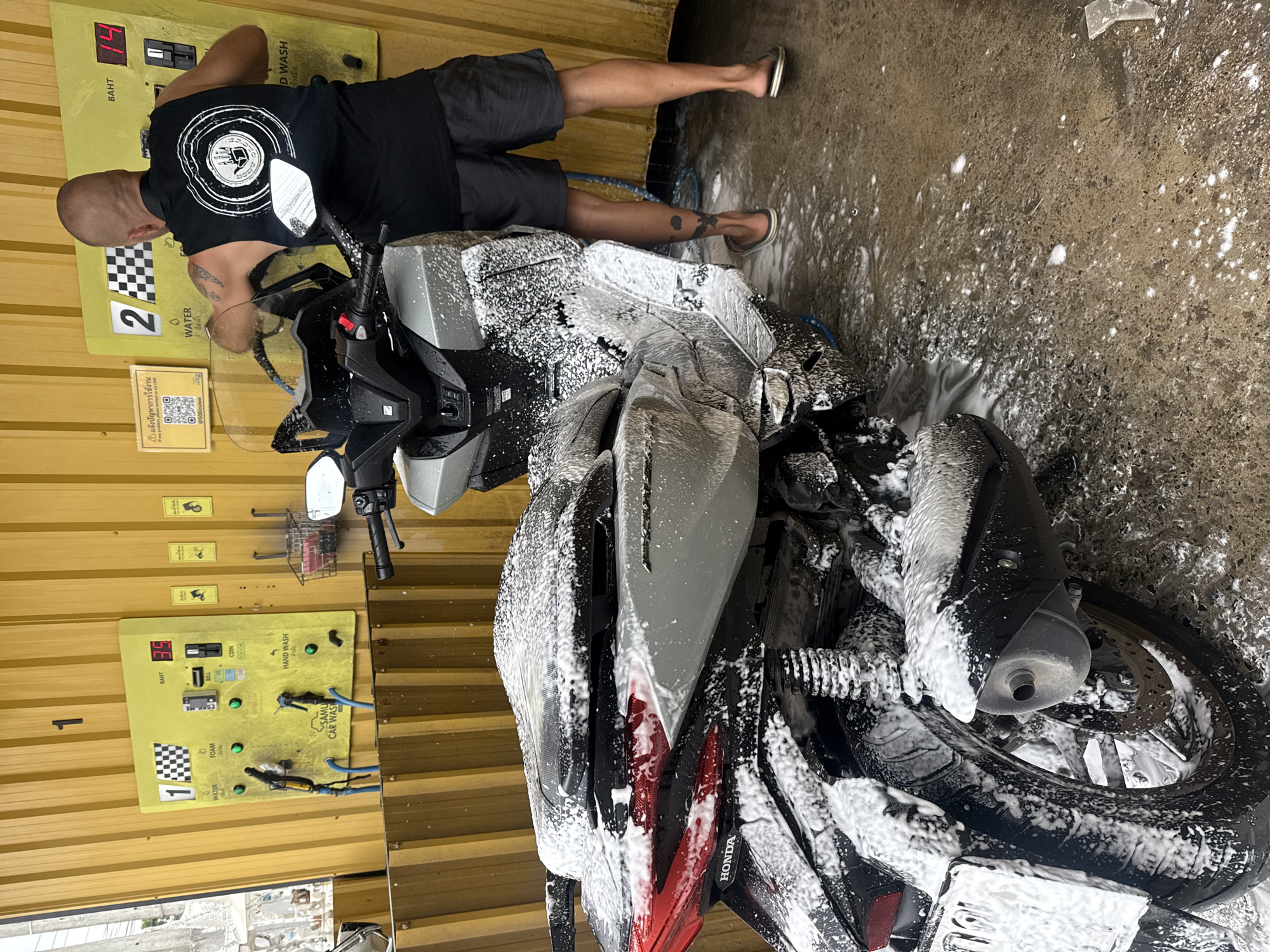 A bald man is washing a silver and black scooter at a self-service car wash in the city of Ko Samui District, Thailand. The car wash is outdoors with a yellow corrugated metal wall in the background. The man, wearing a black sleeveless shirt and dark shorts, is using a foam dispenser on the scooter, which is covered in white foam. He has tattoos on his arms and legs. The scooter is partially disassembled, with parts of the engine and wheels visible. The ground is wet and covered in soap suds. There are several washing stations with control panels that display numbers and instructions in Thai.