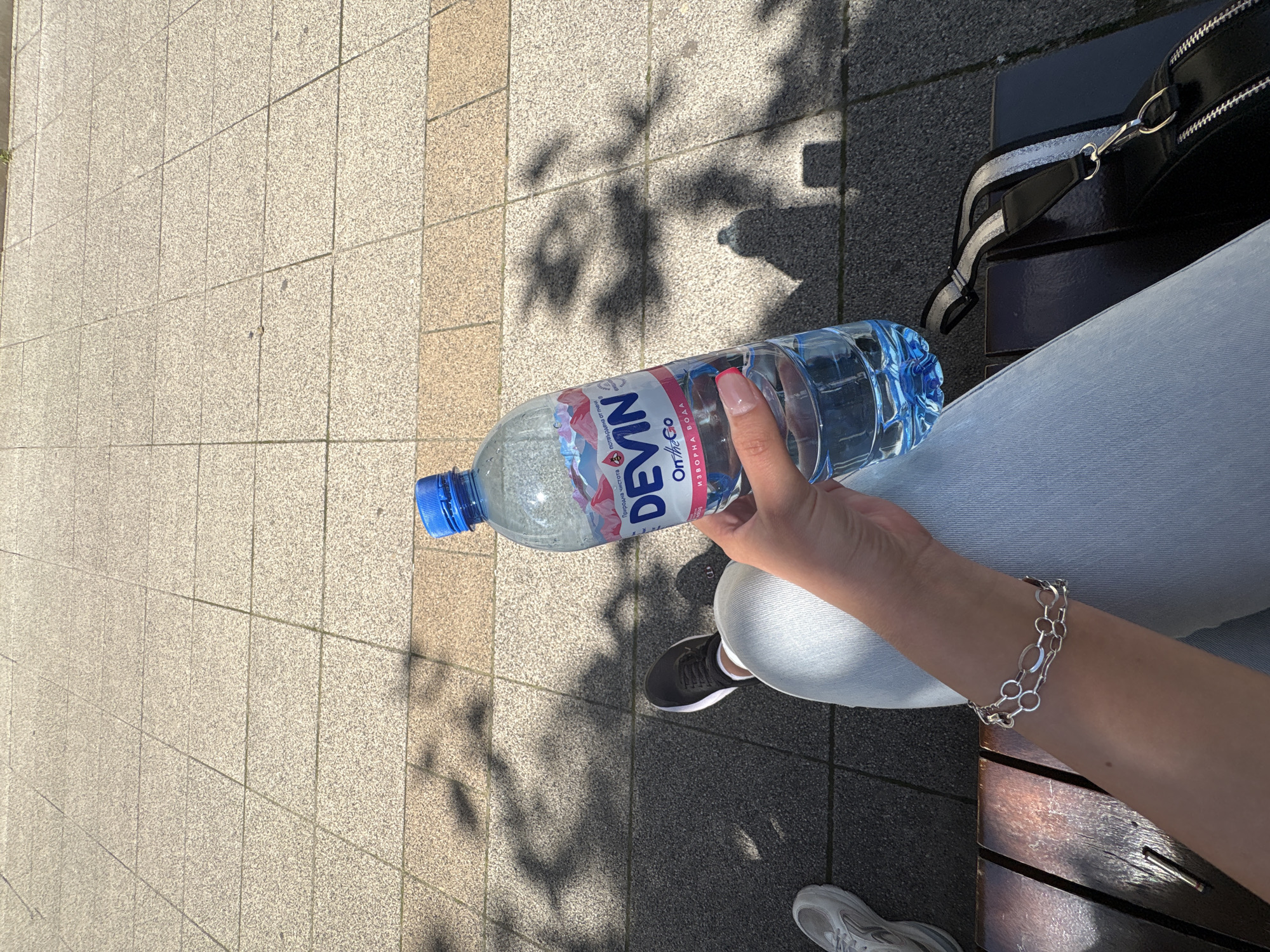 A first-person perspective shot shows a woman's arm holding a bottle of Devin mineral water while sitting on a wooden bench outdoors. The woman is wearing light blue jeans and a silver bracelet on her left wrist. A black handbag with silver zippers rests on the bench next to her. Her black athletic shoe is visible on the ground. The background consists of a tiled walkway and shadows of trees cast on the ground. The scene suggests a casual outdoor setting, possibly a park or public area in Vratsa, Bulgaria.