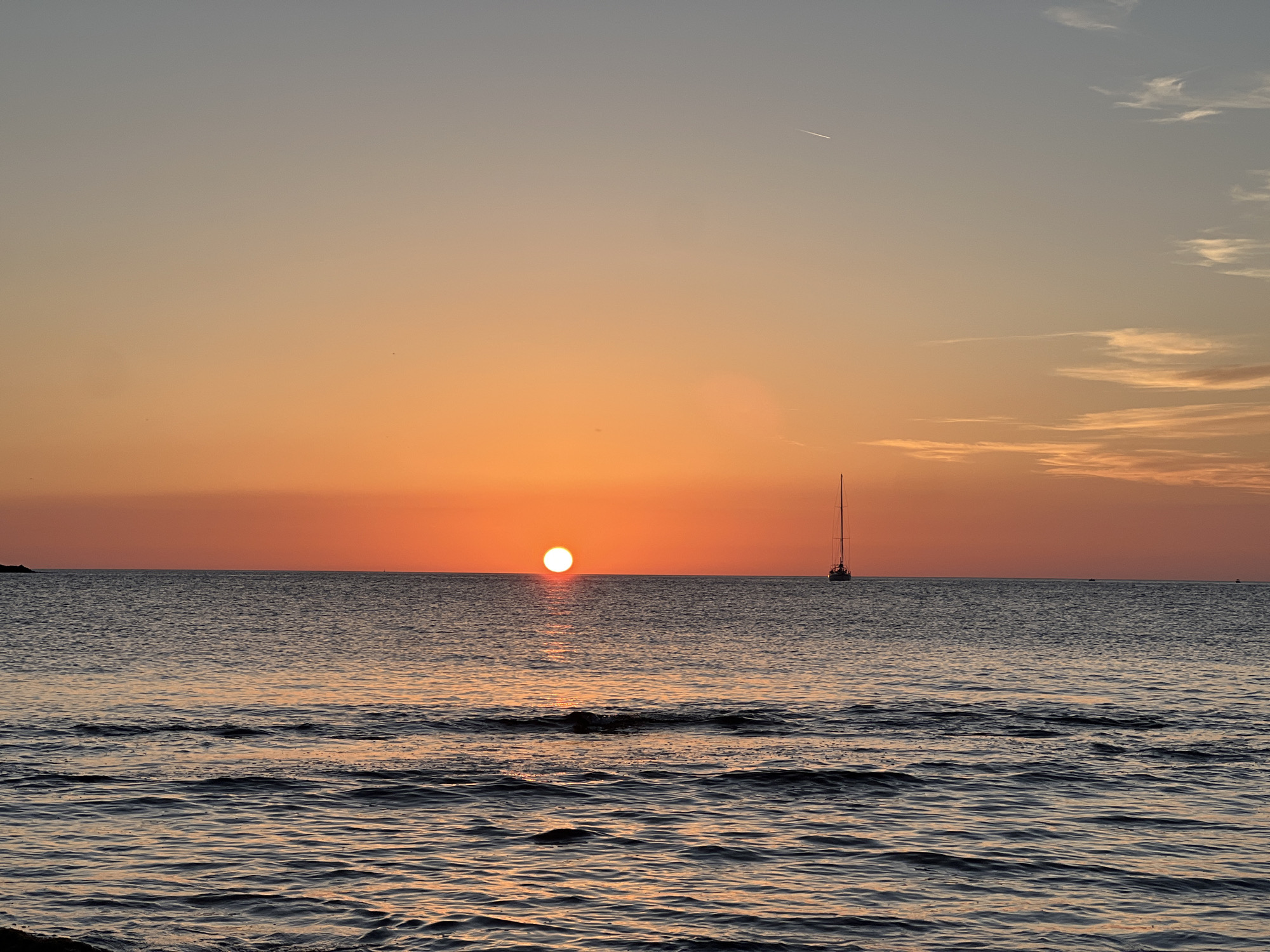 This image depicts a sunset over the sea, with the sun positioned low on the horizon, casting a warm orange glow across the sky. The sea is calm with gentle waves. A solitary sailboat is visible on the right side of the frame, its mast reaching towards the sky. In the distance, on the left side of the frame, a small landmass can be seen. The sky transitions from a pale blue at the top to a vibrant orange near the horizon, with wispy clouds present. The scene is likely a coastal area, given the presence of the sea and the horizon line. While the location is stated to be Palermo, Italy, there are no specific landmarks or architectural features visible to confirm this.