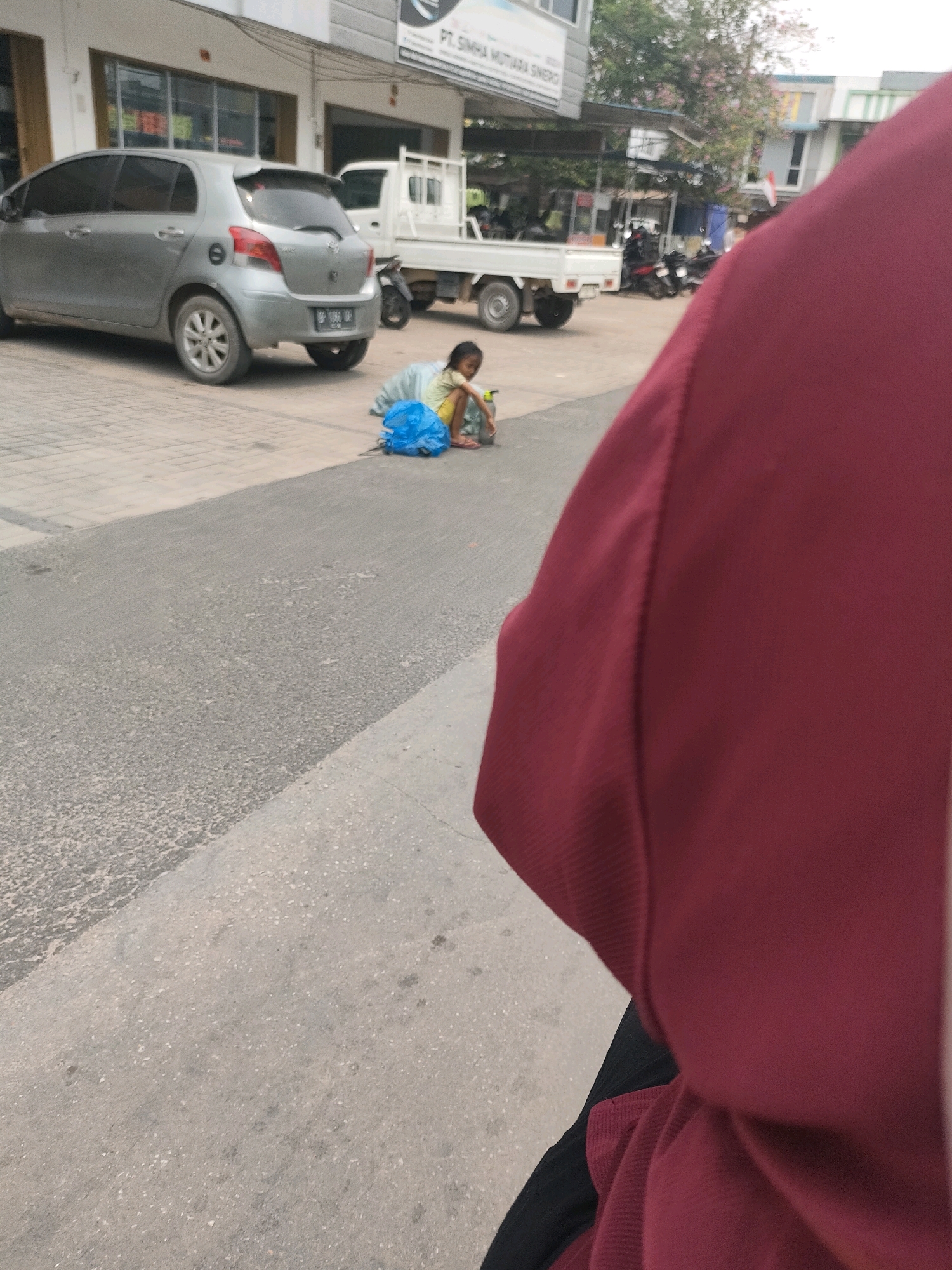In Batam, Indonesia, on a street with a grey asphalt road and a paved sidewalk, a young girl in a light yellow shirt and dark shorts sits on the ground near a large blue plastic bag. She appears to be playing with a small toy car. Behind her, a silver hatchback car is parked on the sidewalk. Further back, a white flatbed truck is visible, along with several motorcycles parked in a lot. The scene is set against a backdrop of commercial buildings. A portion of a person wearing a maroon top is in the foreground, obscuring part of the scene.