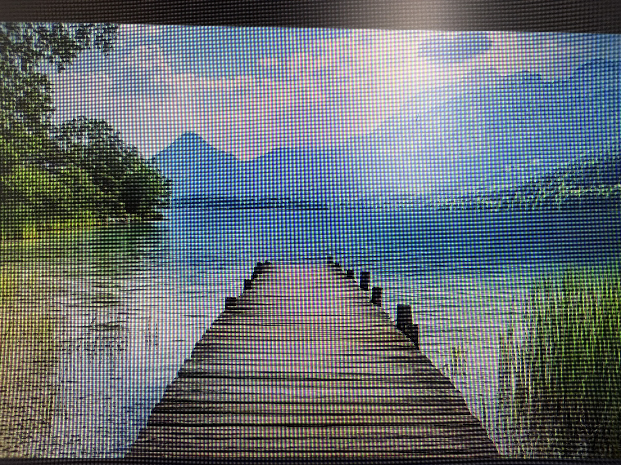 The image depicts a picturesque landscape scene featuring a wooden pier extending into a calm body of water. The pier, constructed from weathered wooden planks, leads the viewer's eye towards distant mountains under a bright sky. Lush green trees line the left bank of the water, while tall reeds grow on the right. The water reflects the sky and surrounding scenery, creating a serene and tranquil atmosphere. The scene is captured from a perspective that emphasizes the length and symmetry of the pier. There are no people or animals visible in the image. The location is not specified as being in the city of Mirnoye, Ukraine.