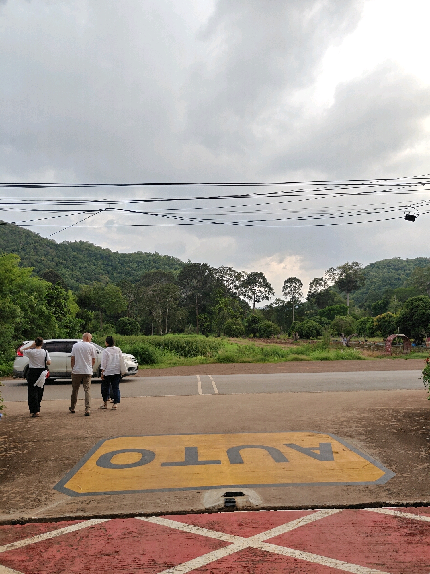 The image depicts an outdoor scene at what appears to be a roadside location in Mu Si, Thailand. In the foreground, a yellow painted area on the concrete ground features dark gray lettering that spells out "AUTO" in reverse, with a gray border. A red concrete surface with white diamond markings is visible at the very bottom of the frame.
Three individuals are standing on the asphalt road, facing away from the viewer. The person on the far left is wearing a white t-shirt and black pants, with their right arm raised to their head. The middle person, a male, is wearing a white t-shirt and tan pants. The person on the right is wearing a white shirt and dark pants. They are standing near a white car parked on the road.
Behind the road, a dense, green forest covers a hilly landscape. The sky above is overcast with gray clouds, with patches of brighter sky visible, suggesting it may be late afternoon or early evening. Power lines are strung across the upper portion of the image. In the distance, on the right side of the frame, some structures and perhaps more vegetation are visible, hinting at a more developed area beyond the natural landscape. The overall impression is a somewhat rural or semi-rural setting with elements of infrastructure like power lines and a roadway.