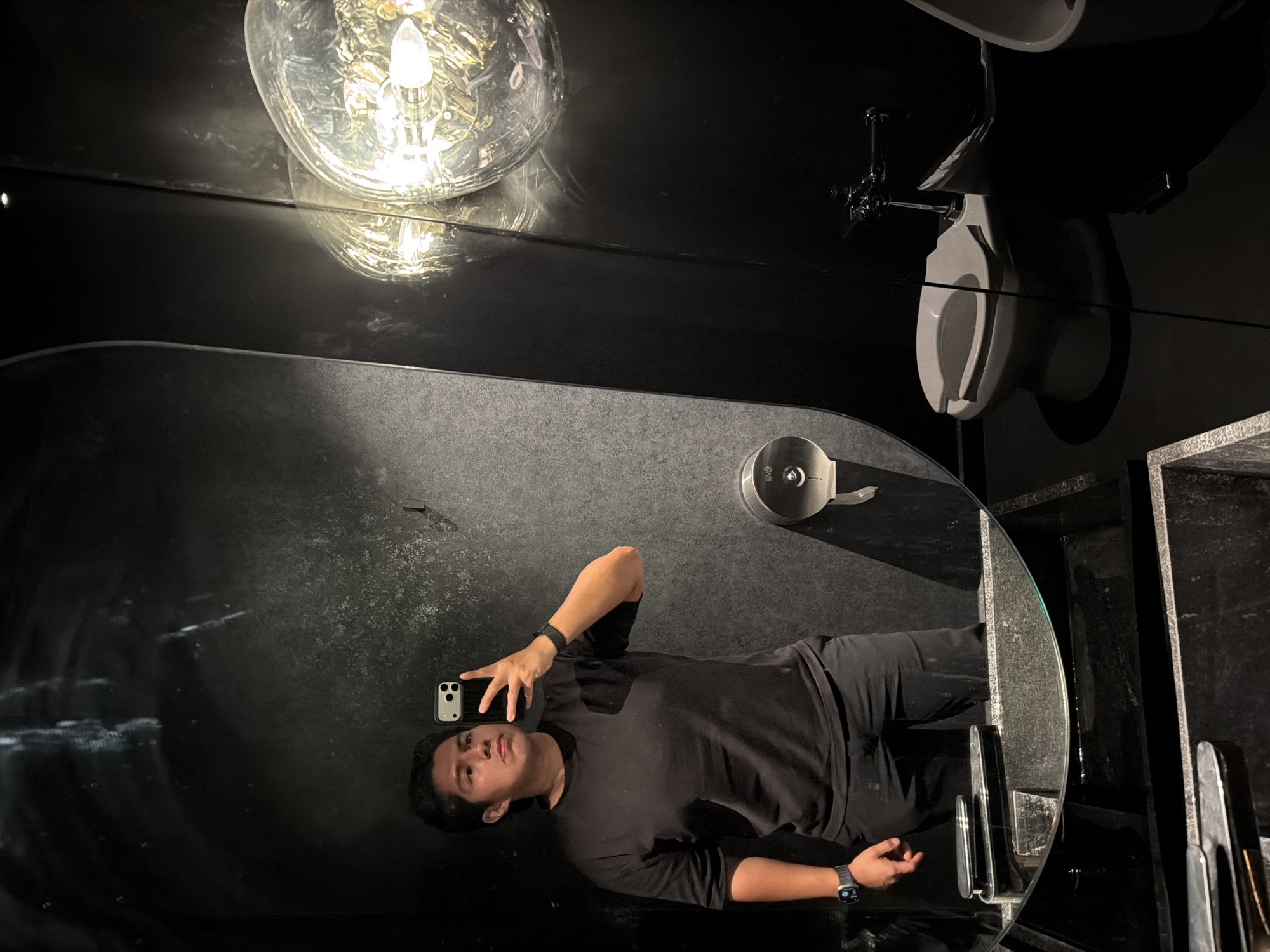 This image, taken in San Bernardino Tlaxcalancingo, Mexico, features a young man taking a selfie in a modern, darkly themed restroom. The man, with dark hair, is wearing a black short-sleeved t-shirt, dark pants, and a dark smartwatch on his left wrist. He holds a black smartphone in his right hand, angled towards himself, and looks directly at the camera with a neutral expression.
He is captured in a reflection on a large, dark, highly polished surface that dominates the scene, likely a mirror or a reflective wall. The room's aesthetic is minimalist and contemporary, with predominantly black or very dark grey surfaces that appear both reflective and textured in different areas.
In the upper right portion of the image, a white toilet is visible, mounted against a dark wall with plumbing fixtures above it. In the reflection, near the man's hip, a round, silver-colored toilet paper dispenser with the text "lavó" is discernible. A notable object is a large, clear glass light fixture with an exposed filament, suspended from the ceiling in the upper left, casting a bright, warm glow that creates prominent reflections on the dark surfaces. The overall lighting is dim, contributing to a sleek, moody ambiance.