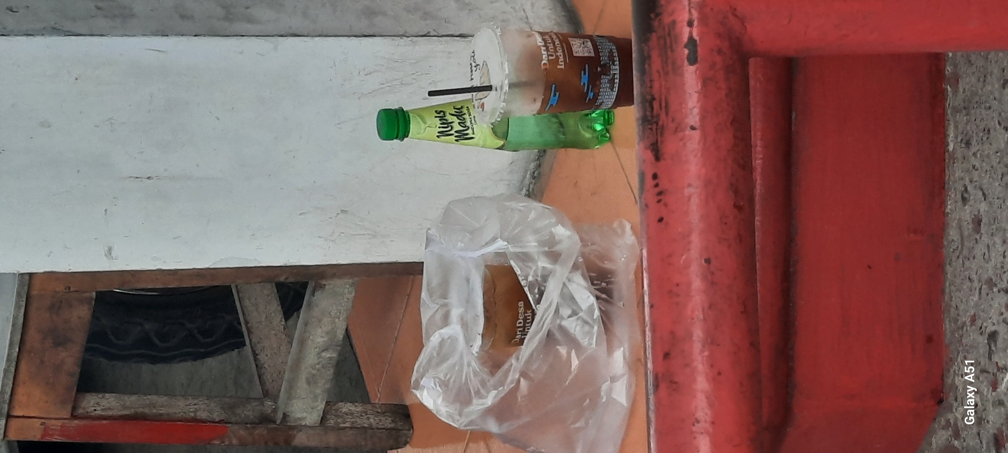 The image depicts an outdoor scene at ground level, likely in a public space in Ploso, Indonesia. In the foreground, a weathered, bright red metal railing runs horizontally across the frame. Behind the railing, the ground is covered with a rough, greyish-white aggregate material.
To the left of the image, a plastic bag contains a drink in a cup, with some text visible on the cup reading "Dari Desa Untuk." Adjacent to the plastic bag, on the right, is a clear plastic cup with a dark brown liquid and a black straw. The lid of this cup has a design with text that appears to say "have you." A green plastic bottle with a green cap stands behind this cup. The bottle has a label with green lettering that reads "Nipis Madu."
In the background, a section of a white wall is visible. To the left of the wall, a rustic wooden structure, possibly part of a table or chair, is present. A black object, possibly a tire or a patterned fabric, is visible within the wooden structure. The overall lighting suggests it is daytime, with diffused light. The scene appears to be a casual, everyday setting.