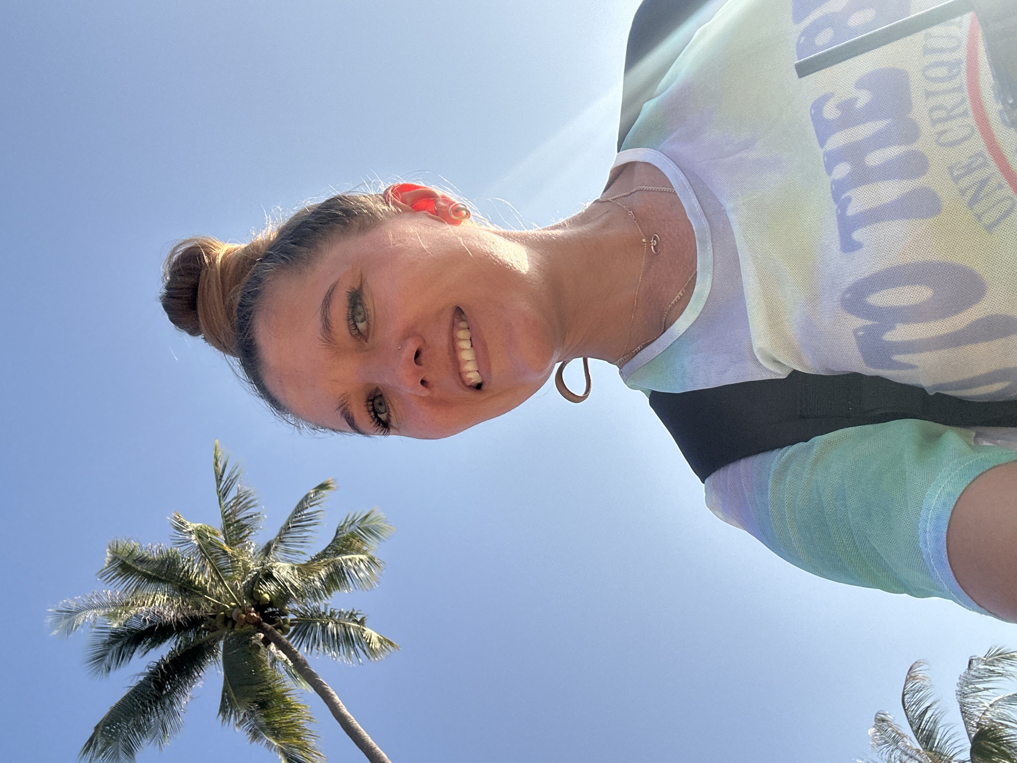 A medium shot shows a young Caucasian woman looking up towards the camera with a smile. She has light brown hair pulled into a high bun. She is wearing a colorful tie-dye shirt with pastel green, blue, and purple hues, and a backpack strap is visible over her left shoulder. A thin gold necklace with a pendant is worn around her neck. Her right earlobe has a small gold earring. In the background, a bright blue sky is visible, with a palm tree extending from the left side of the frame, its fronds reaching towards the top. The sun is shining brightly, casting a lens flare across the image. The scene appears to be outdoors, with the blue sky and palm tree suggesting a tropical location such as Ko Samui District, Thailand.