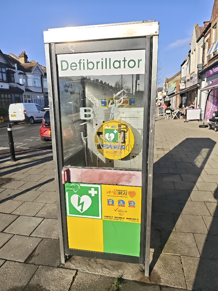 A repurposed, silver-framed public telephone booth, now housing a public access defibrillator, stands on a grey paved sidewalk. The upper panel of the booth displays "Defibrillator" in green text. Behind the scratched and partially stained glass, a yellow circular enclosure contains an Automated External Defibrillator (AED), identifiable by a green heart and lightning bolt symbol, with text indicating "FOR ACCESS CALL 999." The lower section features a green and white symbol of a heart with a lightning bolt, alongside a yellow sign promoting "Community Heart Beat," above solid yellow and green panels. The booth shows signs of wear, including minor paint damage and graffiti on the glass.
To the left of the booth, a black bollard marks the edge of an asphalt street. A white panel van and a red compact car are parked on this street. Beyond, a row of residential houses with pitched roofs, bay windows, and brick facades is visible under a clear blue sky.
To the right, the paved sidewalk extends past commercial buildings. Several individuals are seated at white outdoor tables and chairs in front of a café or similar establishment. A purple banner visible near the shops reads "The Furzedown Project," and a mobility scooter is parked nearby. Shadows are cast across the sidewalk from the right. This scene is located on Mitcham Lane, Streatham, London SW16, United Kingdom.