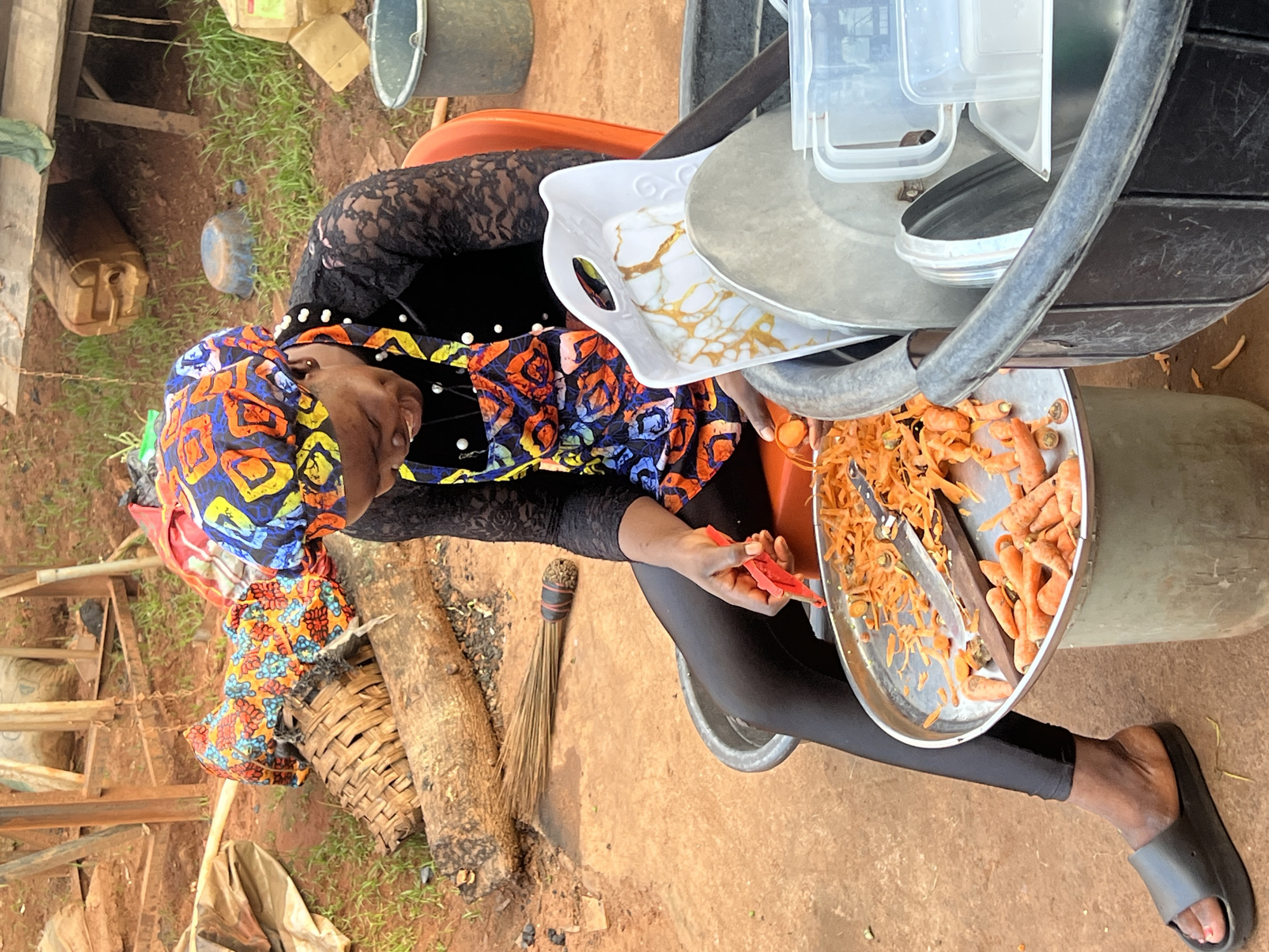 A woman, wearing a black lace top, a colorful headwrap, and black leggings, is seated outdoors in what appears to be a yard in Nkpor, Nigeria. She is holding a red chili pepper in her right hand and appears to be preparing food. In front of her, on a metal tray, are shredded carrots and some whole carrots. A metal grater and a wooden stick are also visible on the tray. To her left, a woven basket sits on the ground. A small broom with a striped handle is leaning against a log next to the basket. In the background, there are several plastic containers, buckets, and wooden structures. The ground is made of reddish-brown soil and patches of green grass are visible. The lighting suggests it is daytime, with no specific indicators of time of day or weather conditions. The woman's expression is not clearly visible, but her focus seems to be on the task at hand.