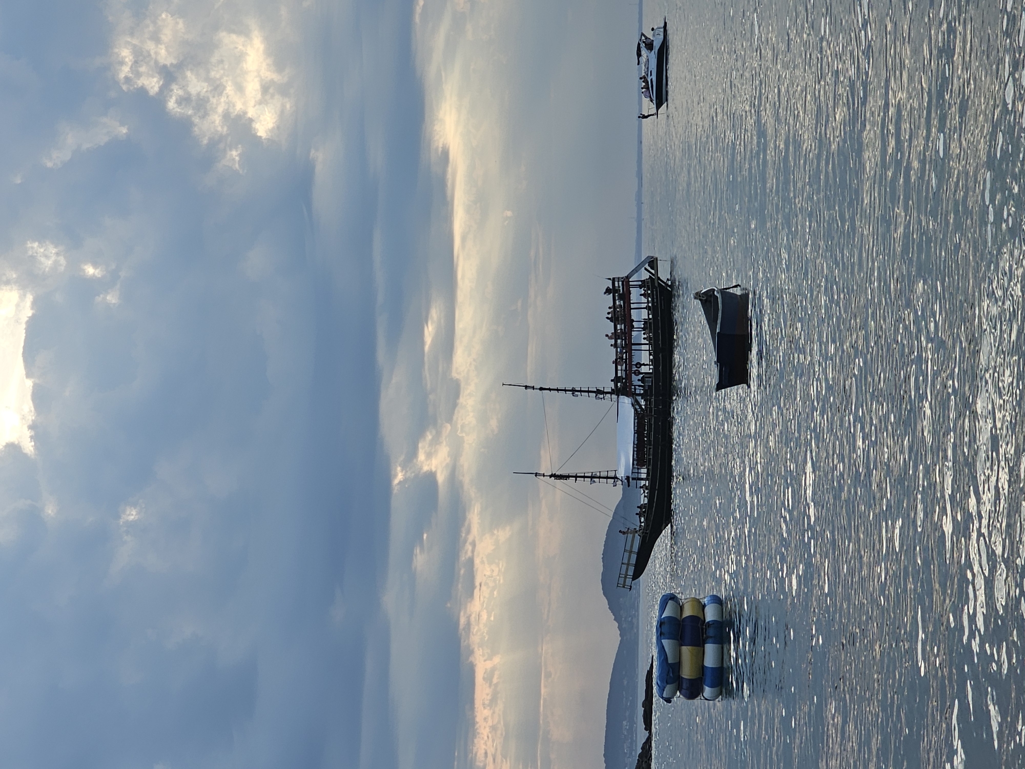 The image depicts a scene in Armação dos Búzios, Brazil at sunset, featuring a body of water with several boats. In the foreground, there are inflatable multicolored floats anchored near the shoreline. A large silhouette of a boat with masts is visible in the water. Further out, a rectangular floating structure appears to be populated with people. In the background, a mountain range is silhouetted against the sunset sky. A smaller motorboat is also present on the water. The sky is filled with clouds tinted with the warm colors of the setting sun, and the water reflects the sky.