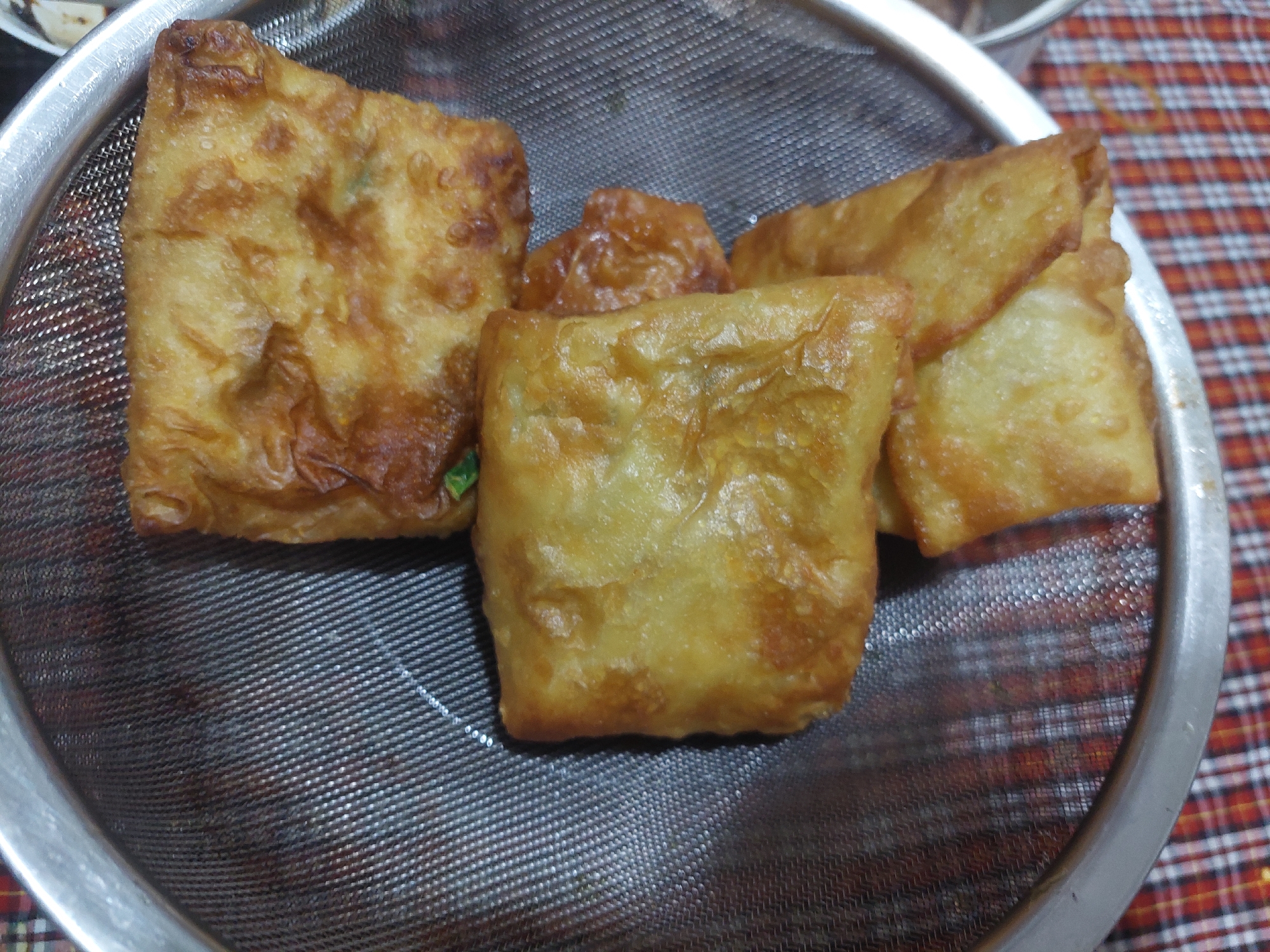 A close-up, overhead shot captures several pieces of fried spring rolls resting on a metal strainer. The spring rolls are golden brown and appear to be filled with a mixture of ingredients. They are arranged in a slightly overlapping manner within the strainer, which is positioned over a red and white checkered surface. The lighting is direct, highlighting the textures and details of the food. The scene appears to be in a domestic setting, possibly a kitchen, in the city of Jelutung, Indonesia.
