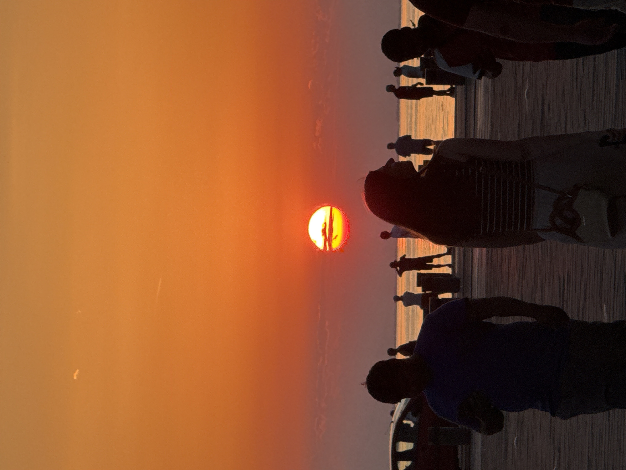 The image depicts a sunset scene at the Molo Audace in Trieste, Italy. Silhouettes of people are visible against the vibrant orange sky, with the sun appearing as a bright, glowing orb partially obscured by a thin layer of clouds. The sun is casting a warm, reddish-orange hue across the sky and the water. Several individuals are seen standing or walking on a wooden boardwalk, their figures outlined by the fading light. In the foreground, two people are the most prominent, with one facing away from the camera and looking towards the sunset, and another in the center of the frame, also facing away. In the background, more silhouetted figures are scattered along the boardwalk and near the water's edge. A car is partially visible on the left side of the image. The overall atmosphere is serene and contemplative, capturing the beauty of a sunset over the Adriatic Sea from a popular waterfront location in Trieste.