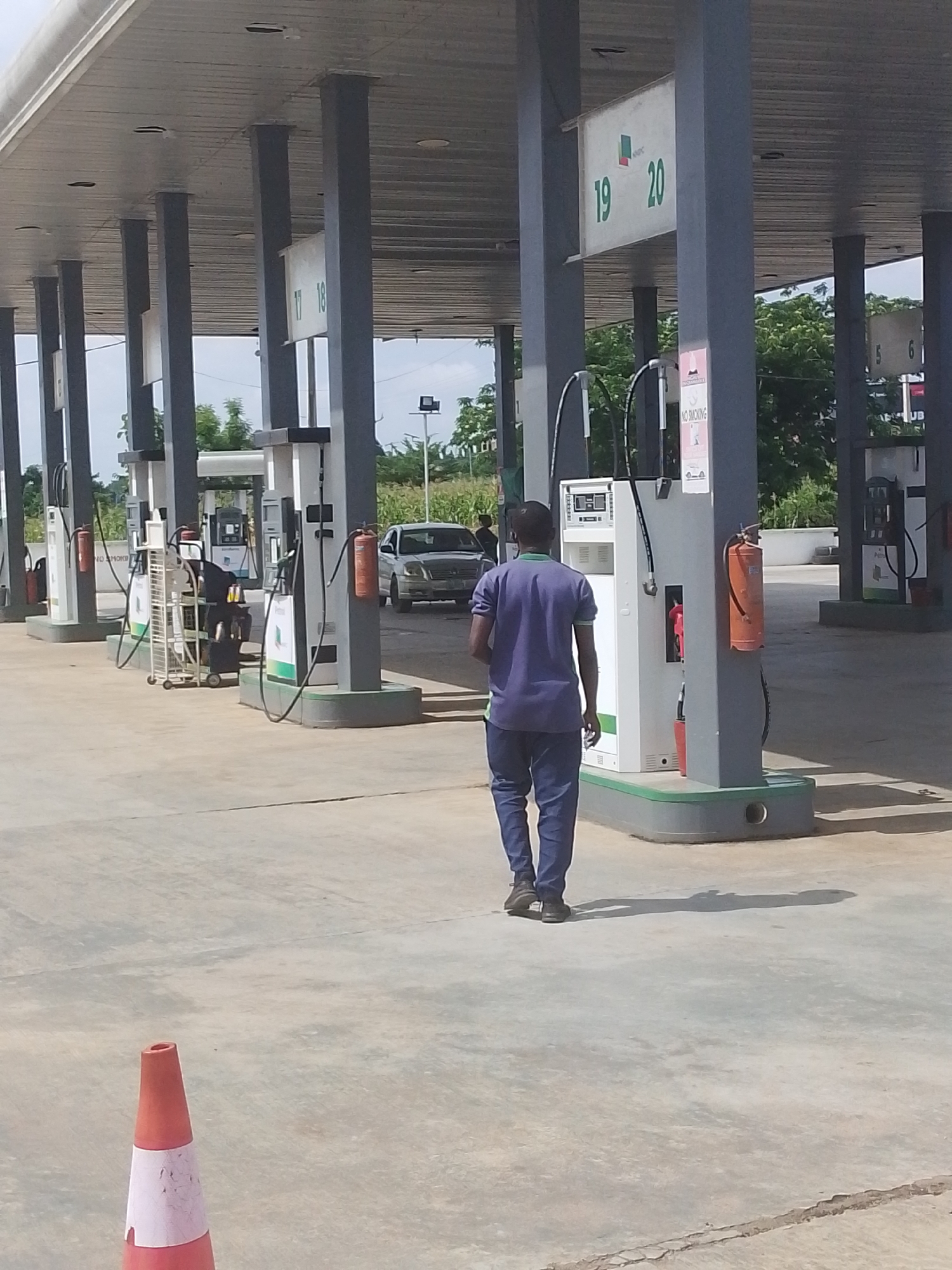 This is a full-shot, eye-level, outdoor, daytime photograph of a man walking away from the camera at a gas station in Abeere, Nigeria. The man is wearing a blue and green polo shirt and blue pants. He is walking on a paved surface. The gas station has a covered canopy supported by gray pillars. Several gas pumps are visible, some with visible branding. An orange fire extinguisher is mounted on one of the pillars. In the background, a silver car is parked at a gas pump, and some greenery and trees are visible beyond the gas station. A red and white traffic cone is in the foreground on the left. The sky is overcast.