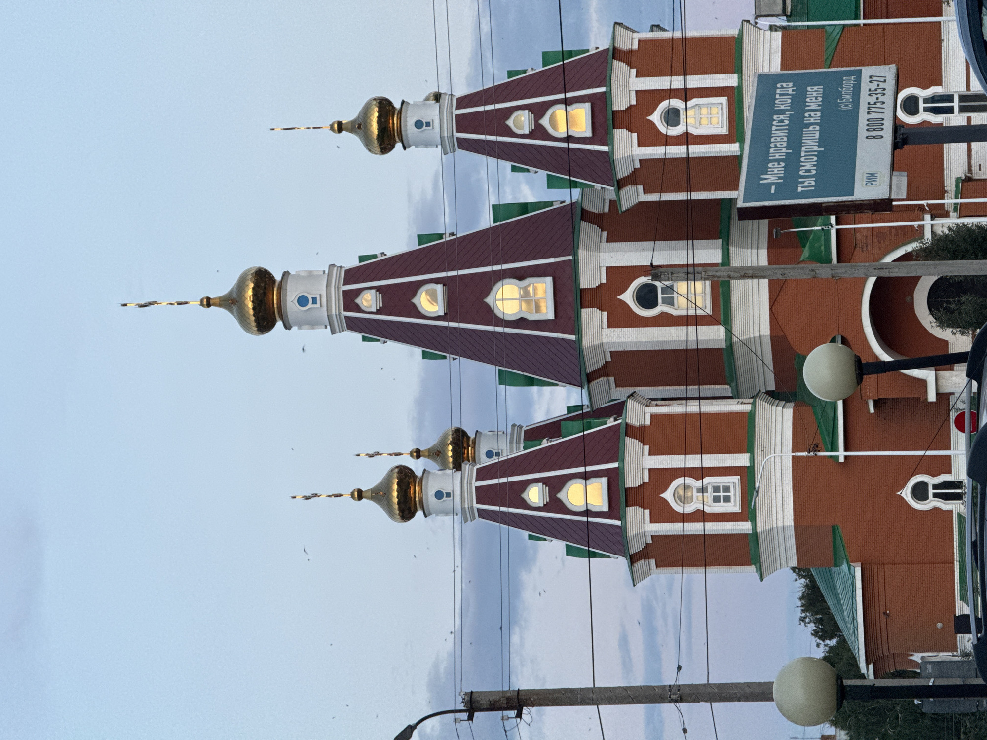 In the city of Otradny, Russia, a brick church with distinctive red and green roofs and golden domes stands prominently against a pale blue, overcast sky. The church features multiple towers topped with crosses, and several windows, some illuminated, dot the facades. In the foreground, power lines and a utility pole are visible, along with a billboard displaying Russian text and a phone number. Two round streetlights stand to the left and right of the frame. A portion of a car is visible at the bottom. The overall scene depicts a religious structure and urban elements under a subdued sky, suggesting it might be dusk or dawn.