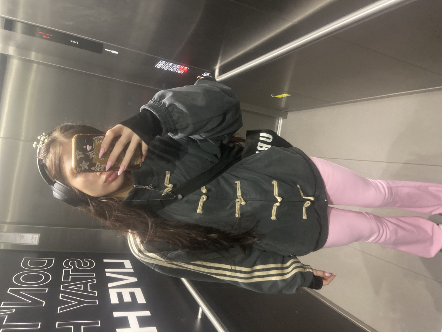 A rotated full-body mirror selfie captures a person standing inside an elevator with metallic, reflective walls. The person is looking into their phone, which is held up to cover their face. They are wearing a dark, possibly padded jacket with cream-colored toggle closures down the front and contrasting gold stripes on the lower left sleeve. Underneath, a black long-sleeved top is visible. The person is also wearing light pink, flared trousers.
Accessories include black over-ear headphones, a small white floral or jeweled tiara-like headband, and a phone case adorned with gold stars and a pink heart decal. The person's nails are long and painted a light pink. A black strap, likely from a bag, crosses their body.
The elevator interior is predominantly grey and reflective, showing reflections of the person and the internal structure. On one of the dark panels, possibly a door or a wall, reversed white text is visible, reading "DON'T STAY" and "LIVE H". Other partially visible, reversed text includes "RU" and "GIVE". The scene appears to be an everyday moment, likely taken in an urban setting like London, United Kingdom.