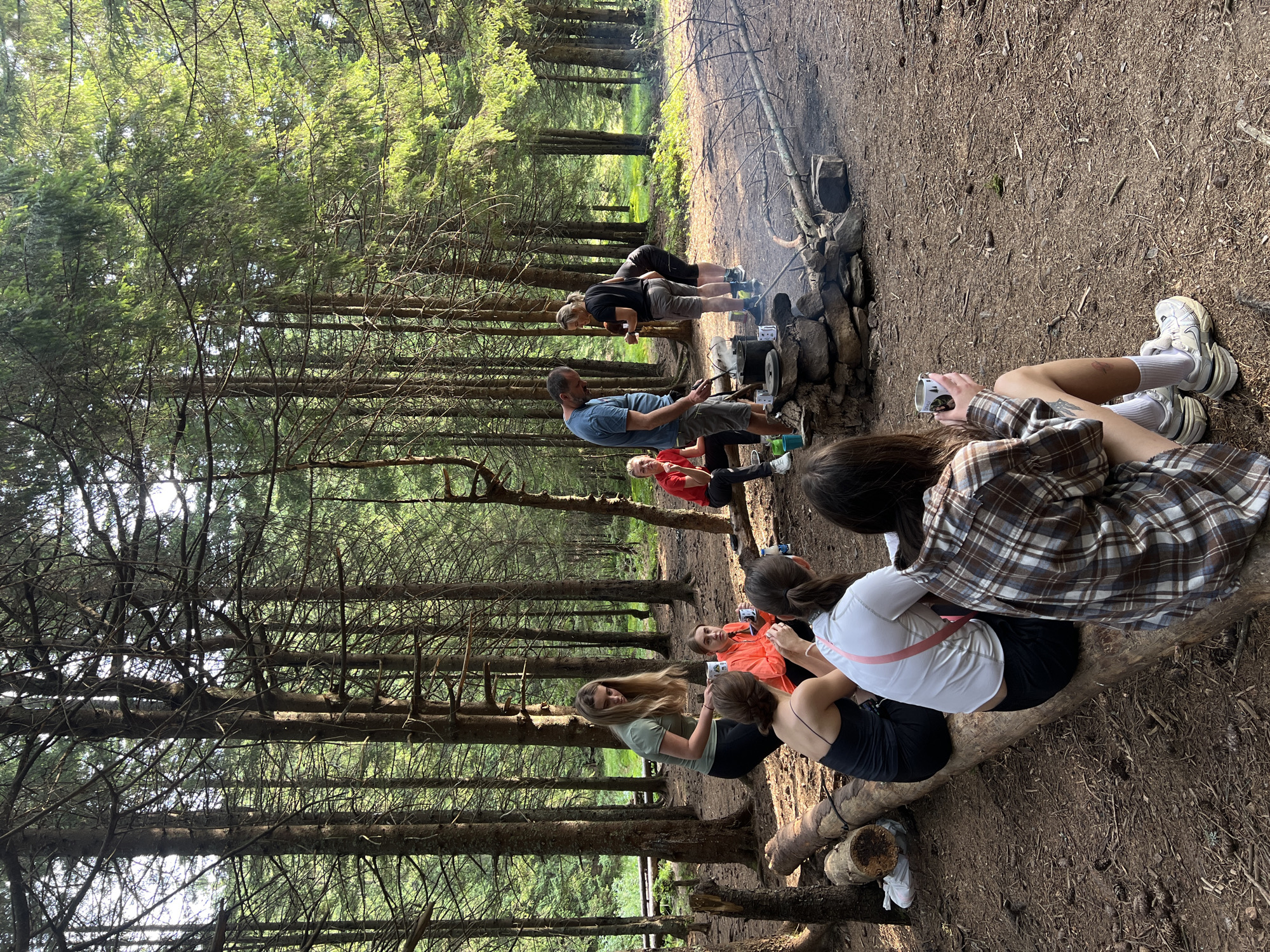 In a forest clearing in Vorokhta, Ukraine, a group of individuals is gathered around a campfire. Several people are sitting on logs or on the ground, while others stand near a makeshift stone fireplace where cooking pots are placed. The environment is characterized by tall coniferous trees, a dirt ground covered with pine needles and fallen branches, and dappled sunlight filtering through the canopy. A few individuals appear to be engaged in cooking or preparing food. The scene depicts an outdoor gathering, possibly a camping or hiking trip, in a natural woodland setting.