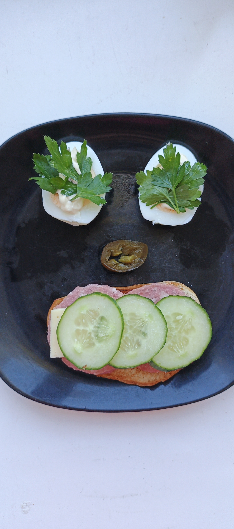 A black plate on a white surface displays a comical arrangement of food resembling a face. Two deviled eggs, garnished with parsley, represent the eyes. A single sliced jalapeño acts as the nose. The mouth is represented by an open-faced sandwich consisting of a slice of bread, cheese, salami, and three cucumber slices. The image appears to be taken in a domestic setting. There is no discernible evidence of the location being Chaikovsky, Russia.