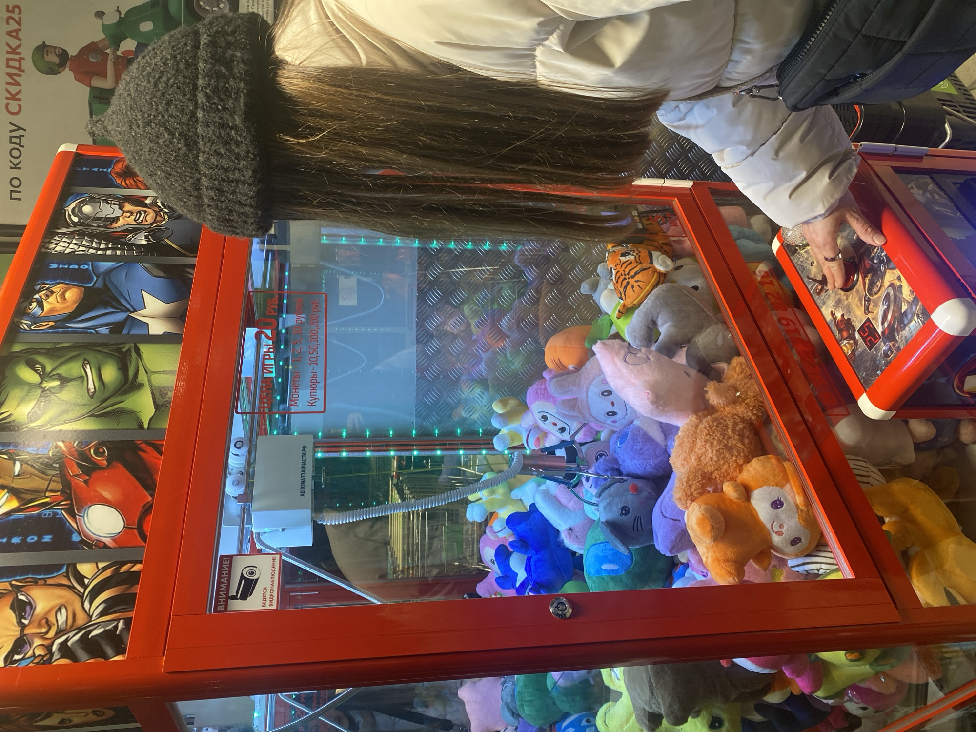 The image depicts a person wearing a gray knit hat and a white puffy jacket, playing a claw machine in Votkinsk, Russia. The machine is filled with plush toys, mostly in pastel colors, including a tiger and several animals with embroidered faces. The frame of the machine is red and features comic book superhero graphics. The control panel is also red with superhero-themed buttons and decorations. There is Russian text on the machine, indicating it accepts coins and banknotes, and a warning sign stating video surveillance is in operation. The person's hand is visible on the control panel, apparently manipulating the claw.