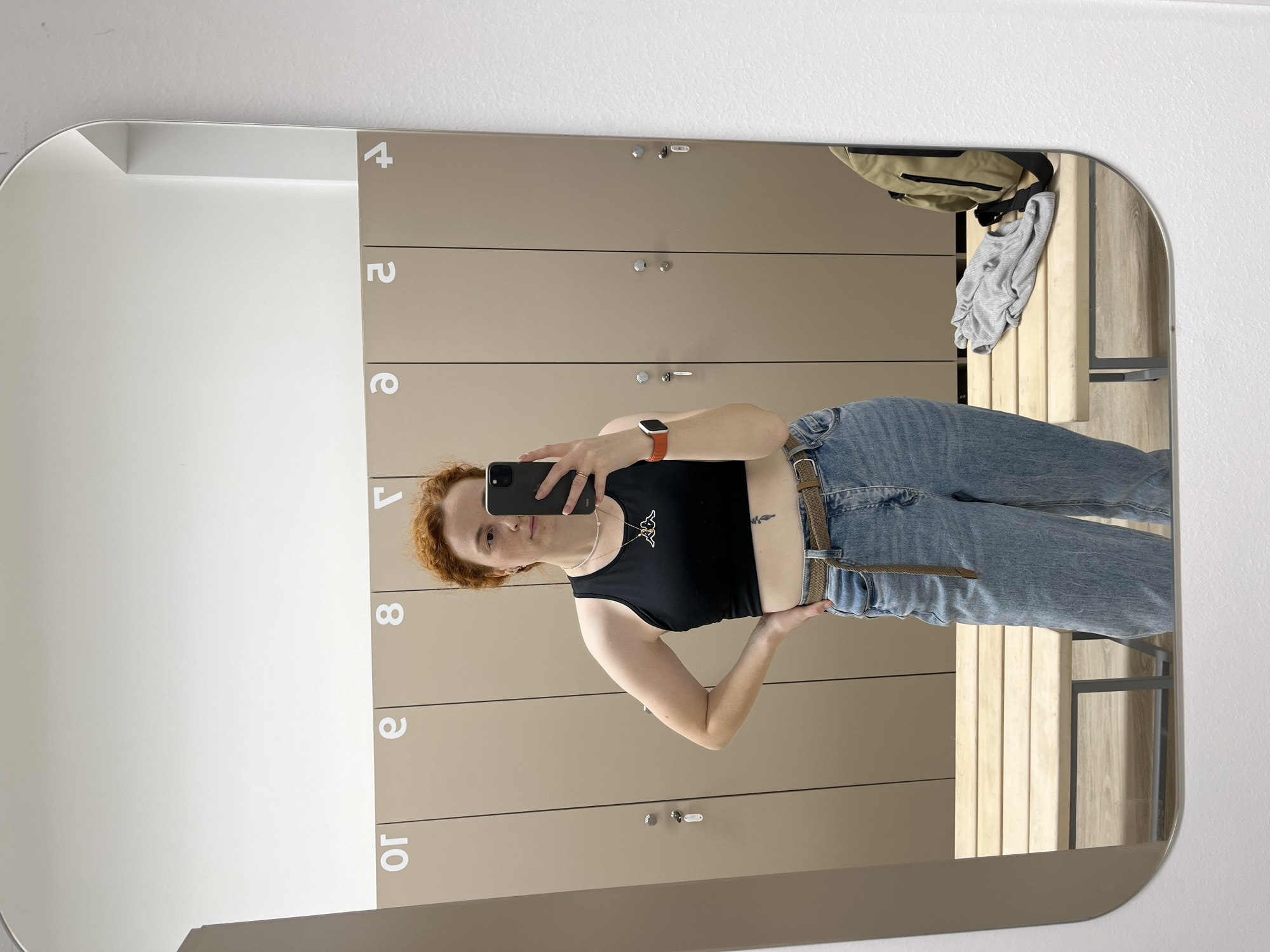 A person is visible in a mirrored reflection, seemingly in a locker room or changing area, likely in the city of Chaikovsky, Russia, based on possible context. The individual is holding a mobile phone, apparently taking a photograph of themselves. The attire includes a dark-colored top, blue jeans, and a belt. The reflection reveals a row of numbered lockers behind the person. A bench and various items, possibly clothing, are visible in the upper right of the mirror's reflection. The mirror is mounted on a wall next to a white vertical surface.