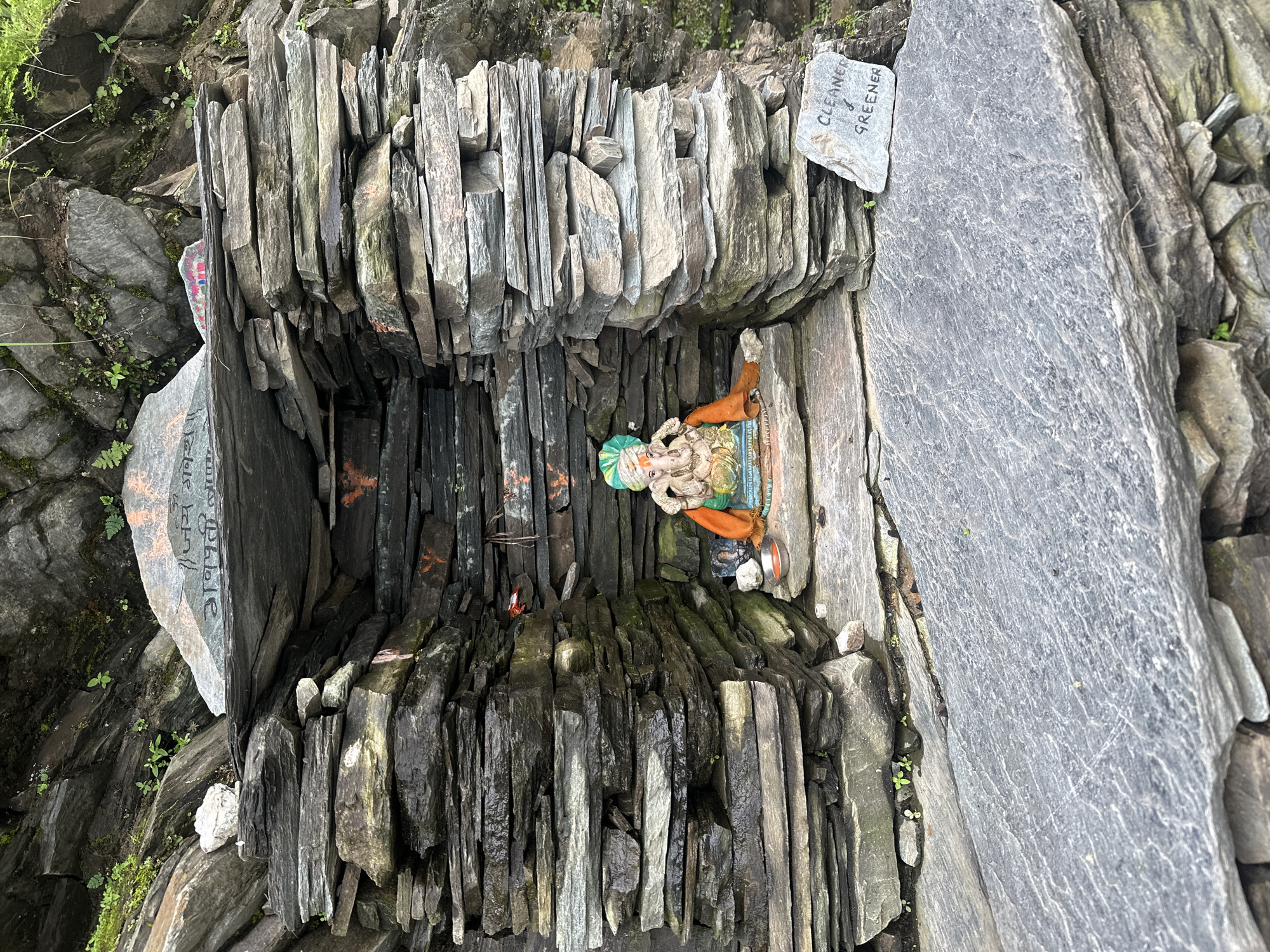 In Bhagsu, India, a small shrine is constructed from stacked, dark gray slate stones. The shrine is built into a rocky hillside, with greenery visible in the background. A statue of the Hindu deity Ganesha, adorned with an orange cloth and a green turban, is placed inside the shrine. To the right of the shrine, a small, light gray stone with the inscription "CLEANER & GREENER" is visible. The overall scene depicts a simple, naturalistic shrine in a mountainous environment.