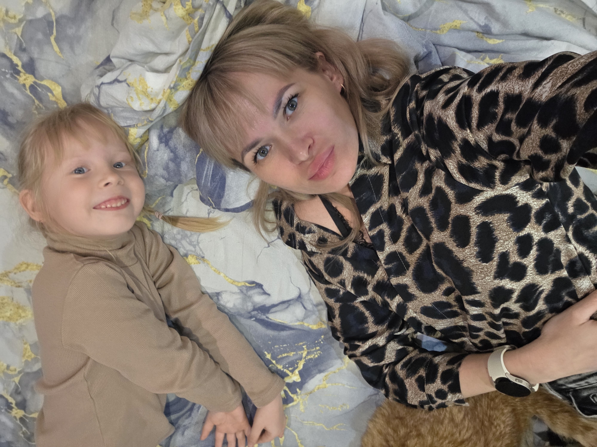 An overhead shot taken in Chaikovsky, Russia, captures an adult woman and a young child lying side-by-side on a bed. The bed is covered with a distinct blue, grey, and gold abstract patterned bedspread.
The adult woman, positioned on the right side of the frame, has blonde hair with bangs and striking blue-grey eyes. She wears a long-sleeved top featuring a prominent brown and black leopard print. A white or light-colored smartwatch with a dark screen is visible on her left wrist. Her expression is relaxed and a bit playful, with a slight pout.
To her left, a young child, likely a girl, with blonde hair and bright blue eyes, smiles broadly at the camera, showing her teeth. She is dressed in a plain, ribbed beige turtleneck top. Both individuals appear comfortable and happy, suggesting a candid moment of leisure or affection.
The setting is an indoor space, presumably a bedroom. Partially visible in the bottom right corner is a patch of brown fur, indicating the presence of a pet, possibly a cat or dog, lying close by. There is no visible text in the image.