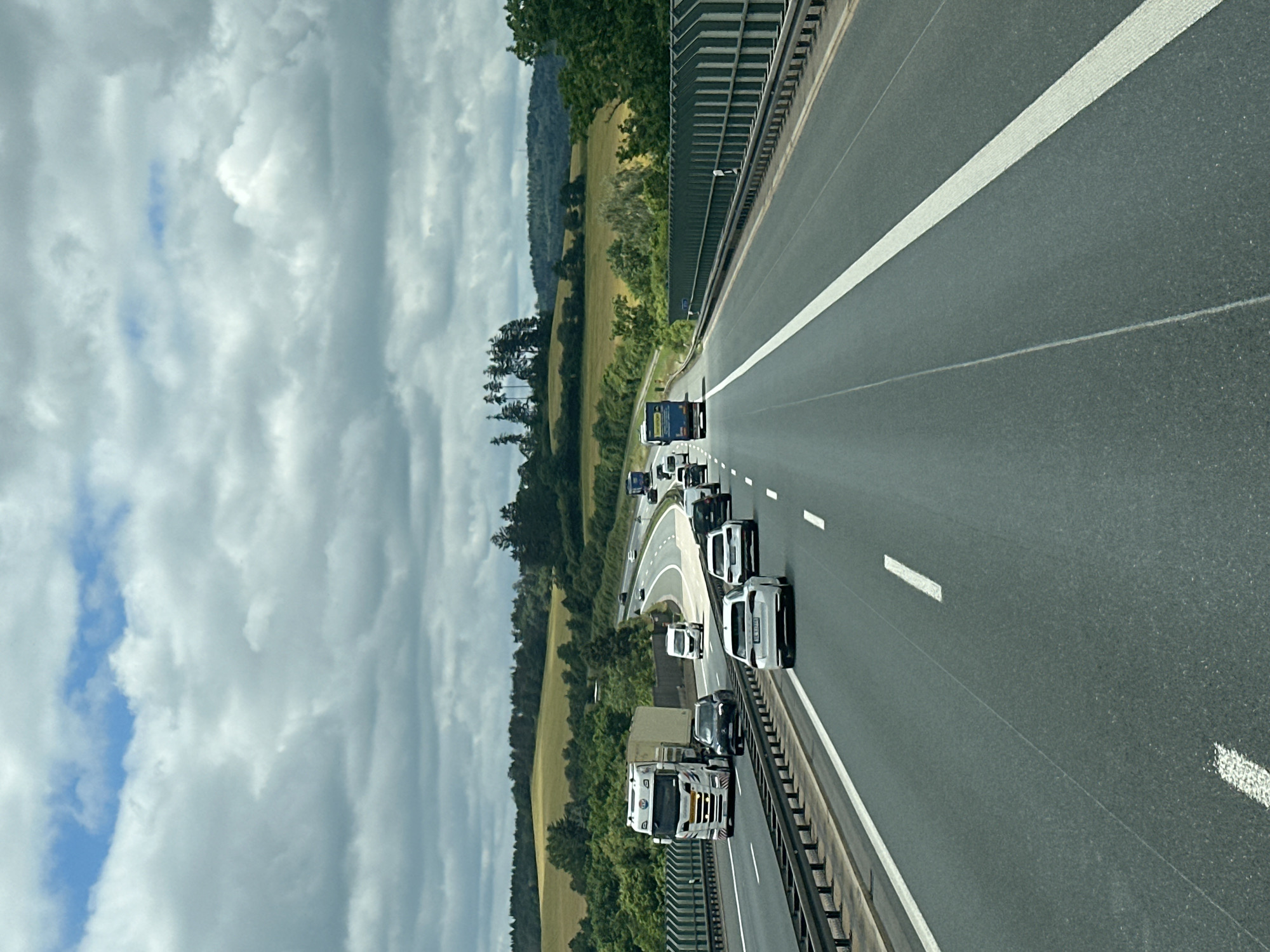 A daytime, eye-level, vertical shot shows a multi-lane highway on a cloudy day. The highway is asphalt, with white lane markers. Guardrails are visible on both sides of the road. Numerous vehicles, including cars, trucks, and vans, are traveling on the highway. The surrounding environment consists of green hills and trees, suggesting a rural or semi-rural location. The sky is filled with a mix of white and gray clouds, with patches of blue sky visible. The specific location is on the A72 highway near Weischlitz, Germany.
