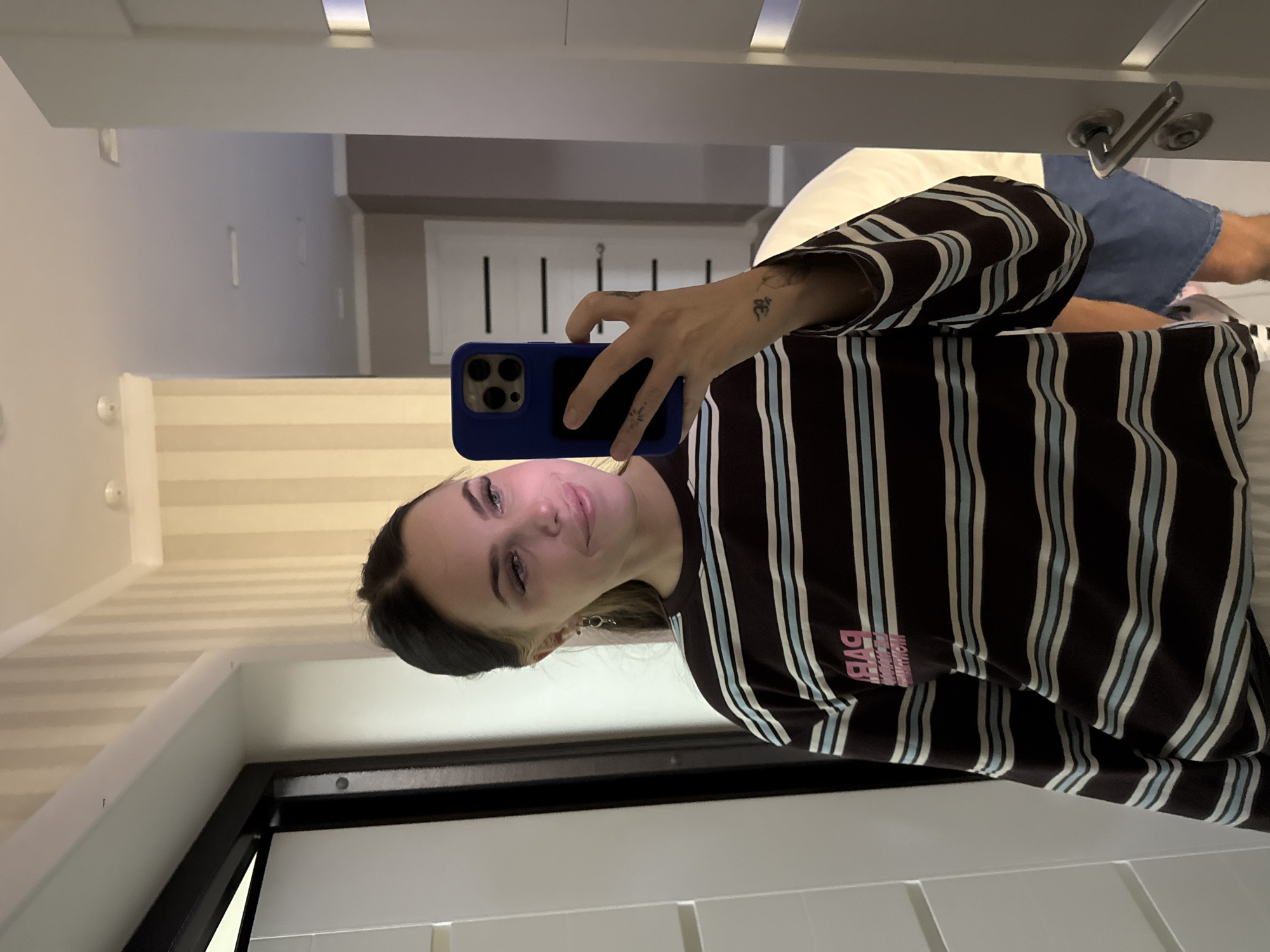 A young Caucasian woman with dark hair pulled back into a ponytail is taking a mirror selfie in a hallway. She is wearing a long-sleeved striped shirt in brown, blue, and white. She is holding a blue smartphone in her right hand, which is angled to capture her reflection. She has a slight smile on her face and is looking at her reflection in the mirror. The hallway has striped wallpaper in cream and beige tones and appears to be a residential setting. A door is visible in the reflection to the right, and a portion of another door or wall is visible on the left. There is a visible light fixture on the ceiling. A person wearing a white shirt and blue shorts is partially visible in the reflection behind the woman. The scene is consistent with an interior residential setting in a city like Chelyabinsk, Russia.