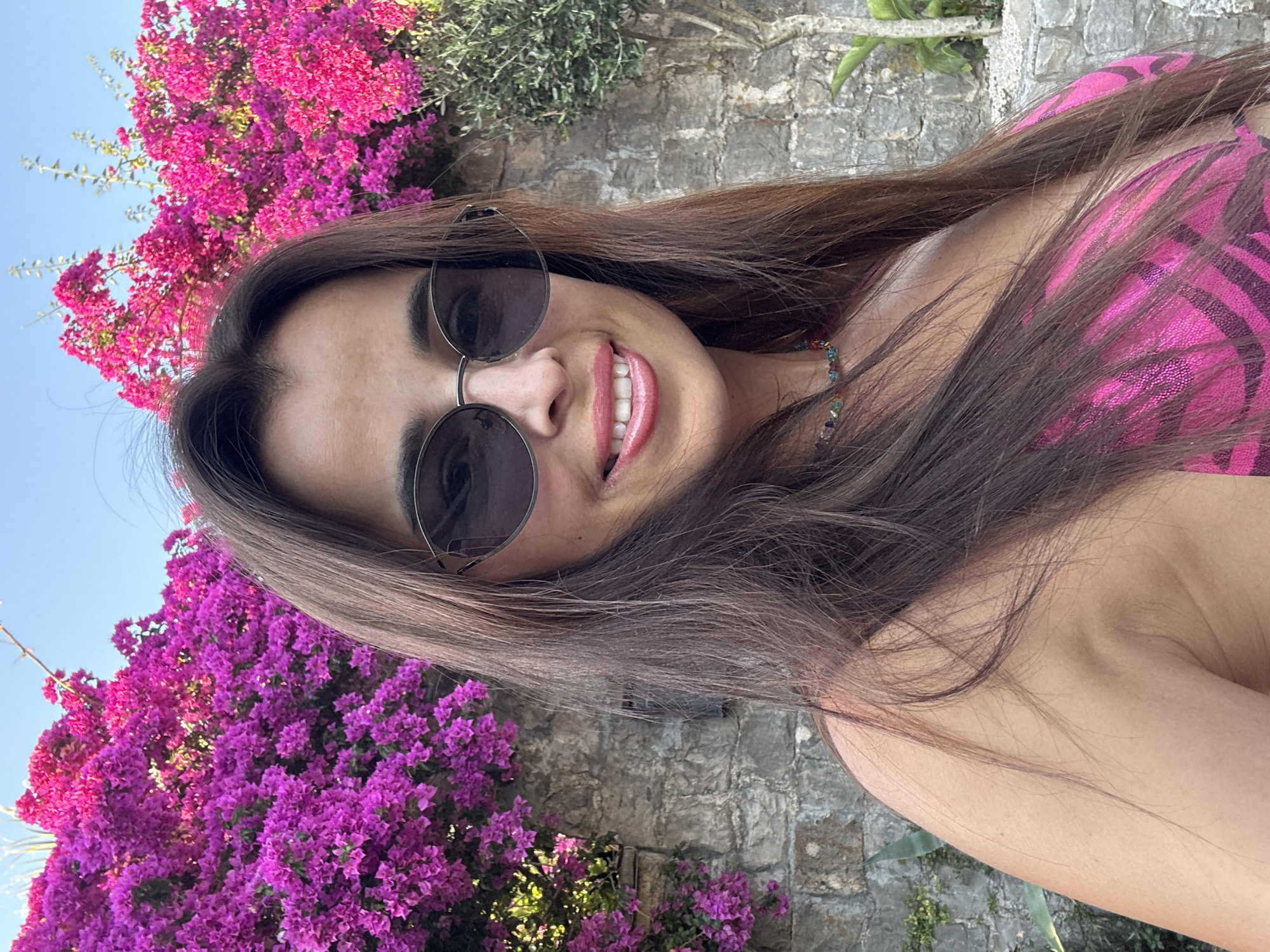 The image shows a woman in a selfie. She is wearing sunglasses and a pink and black patterned top. Her hair is long and brown. She is smiling and has on pink lipstick. She is outdoors, with vibrant pink bougainvillea flowers in the foreground and background. A stone wall is visible behind her. The lighting suggests it is daytime, possibly late afternoon due to the warm glow. The overall mood appears happy and relaxed. There is no visible text in the image.
