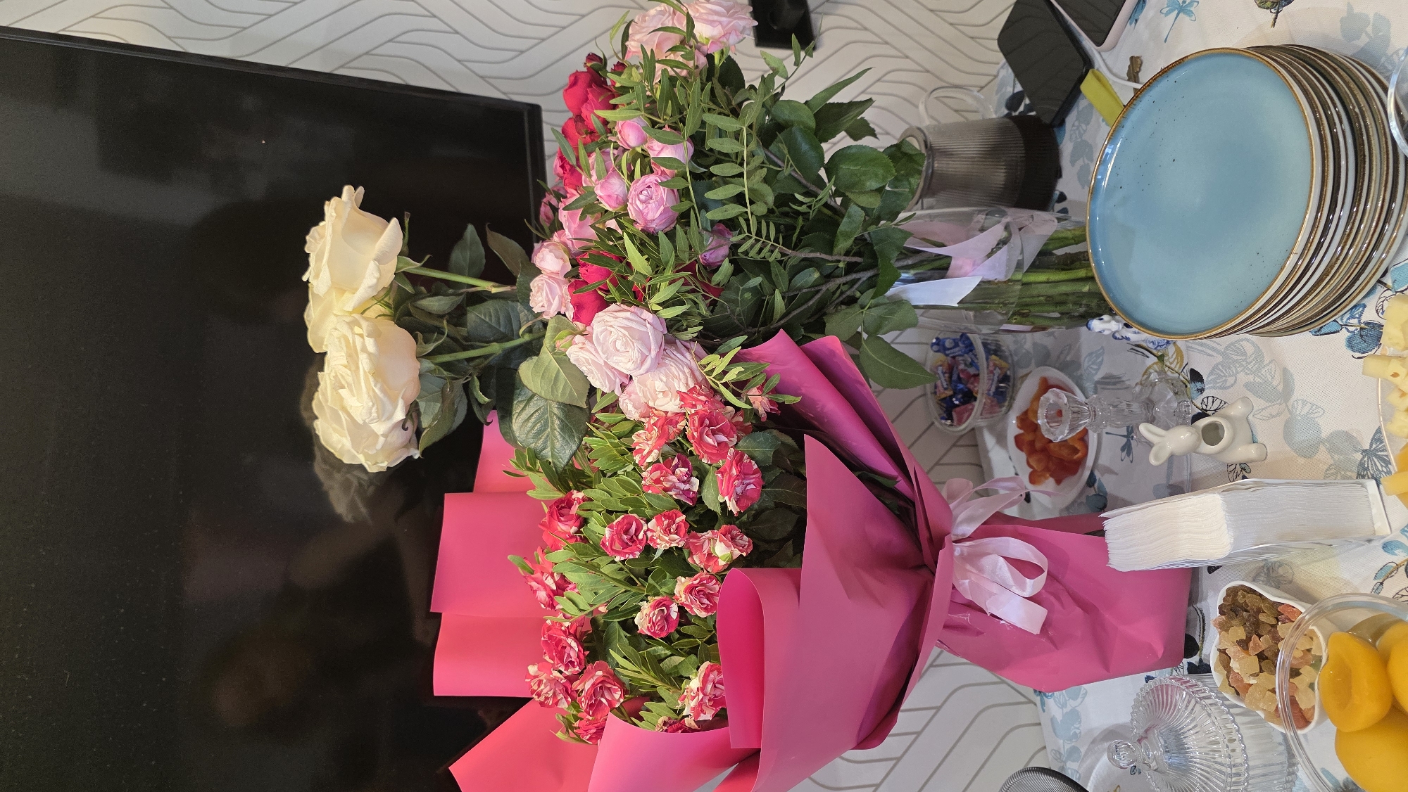 A still life photograph captures a colorful display of flowers and food items on a table. Two bouquets of roses are prominently featured: one with cream-colored roses and another with a vibrant mix of pink and red roses, both wrapped in bright pink paper and tied with white ribbons.
On the table, to the right of the flowers, is a stack of blue-rimmed plates, a small white ceramic rabbit figurine, a clear glass candle holder with sliced red peppers, and a bowl filled with various dried fruits and jellied candies. A glass bowl containing sliced peaches is also visible. Further to the right, a portion of cheese and yellow fruit can be seen.
In the background, a black surface, likely a television screen, reflects some of the light and the bouquet. The wall behind is decorated with a patterned wallpaper. The overall setting suggests a festive occasion or a special presentation of gifts and refreshments. The lighting indicates it is daytime. There is no visible text in the image. The scene is set in Ufa, Russia.