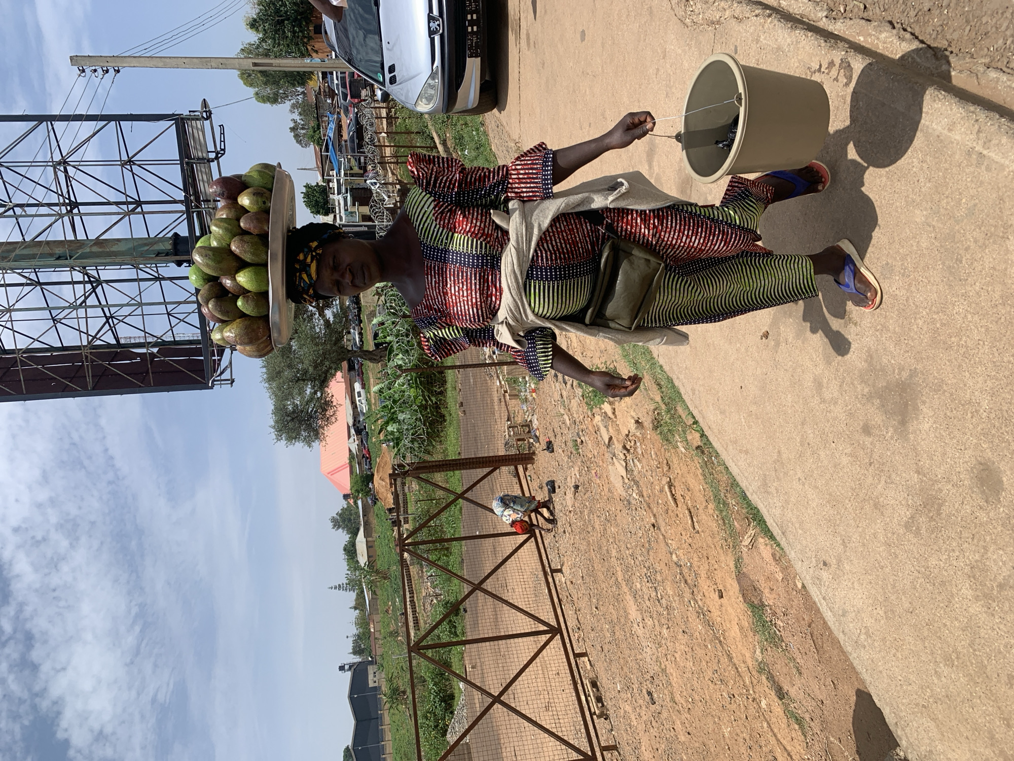 A woman walks on a sidewalk next to a dirt road with fenced plots in Bukuru, Nigeria. She carries a tray of avocados balanced on her head and holds a bucket by its string. The woman wears a colorful patterned dress, a light-colored wrap, and sandals. A large metal structure stands next to the road. Cars are parked in the background. A smaller figure is visible behind the fence.