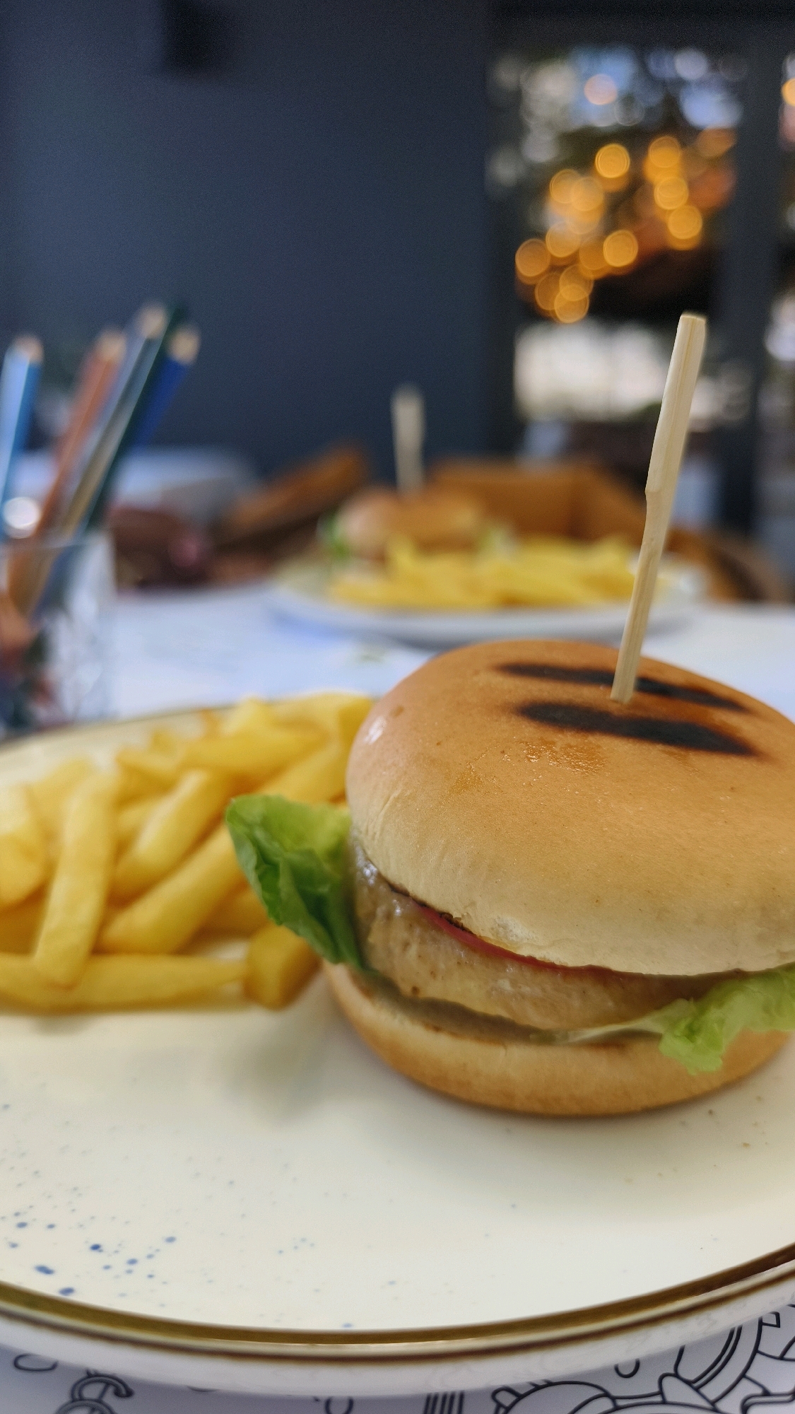 A close-up, shallow depth of field shot captures a mini burger and fries on a white plate with a gold rim. The burger, speared with a wooden pick, is in the foreground, slightly to the right, featuring a grilled patty, lettuce, and bun. To the left of the burger are golden french fries. The background is blurred, showing hints of other food on plates and an out-of-focus, dimly lit environment with bokeh lights. There are also colored pencils in a holder on the left side of the frame, suggesting a casual or artistic setting. The scene appears to be indoors, possibly in a restaurant or cafe in Vienna, Austria. No people are visible in the frame.