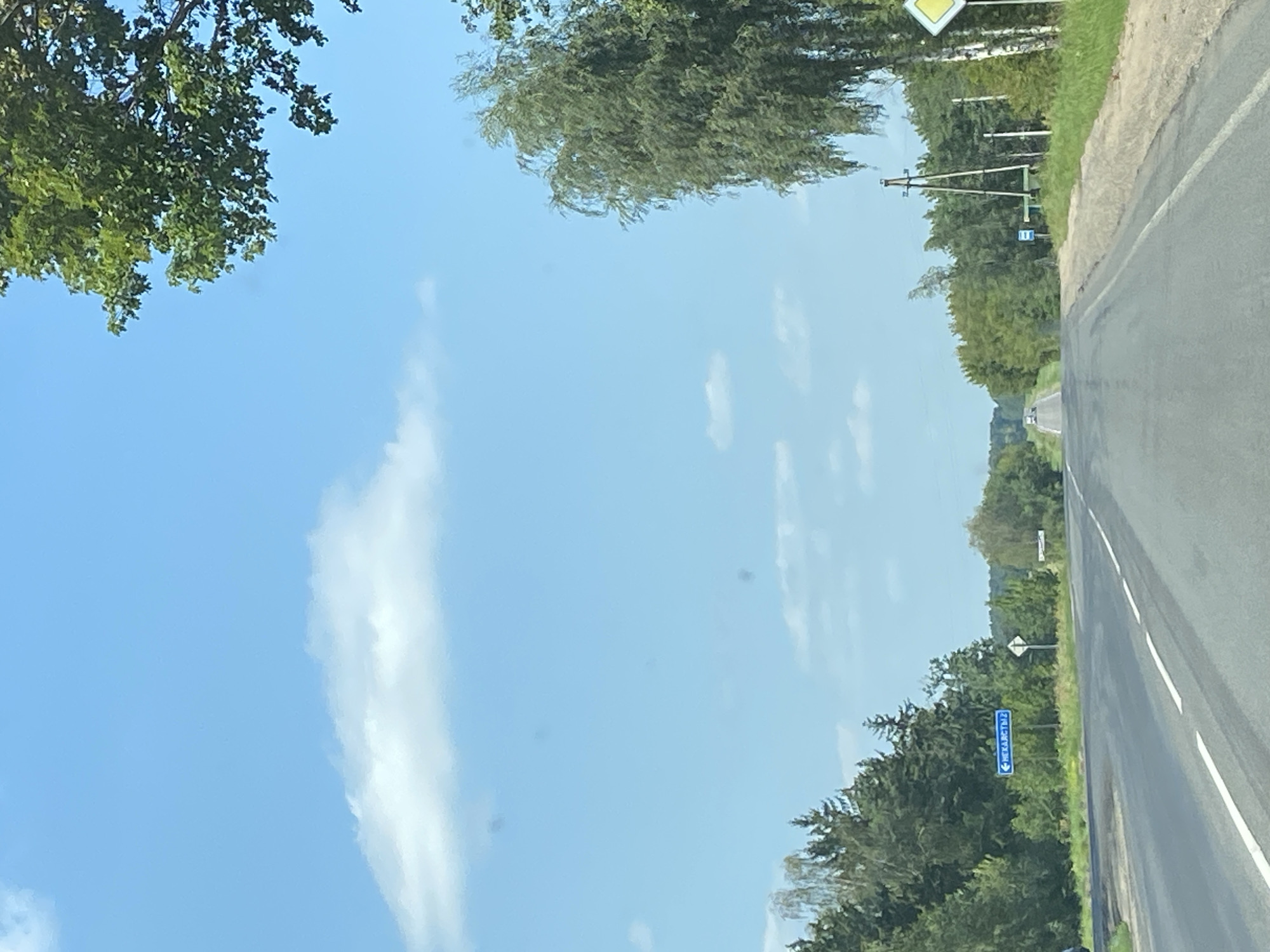 The image depicts a clear, blue sky with scattered white clouds over a rural landscape. A paved road curves to the left, bordered by trees and a grassy verge. On the left side of the road, a blue sign with white Cyrillic text, likely indicating a location name, is visible. Further down the road, in the distance, two white cars are driving away from the camera. On the right side of the road, there is a yellow diamond-shaped sign with a black border, and a tall pole with some wires. The scene appears to be a serene, sunny day in the countryside, possibly in or near Матыкальскі сельскі Савет, Belarus.