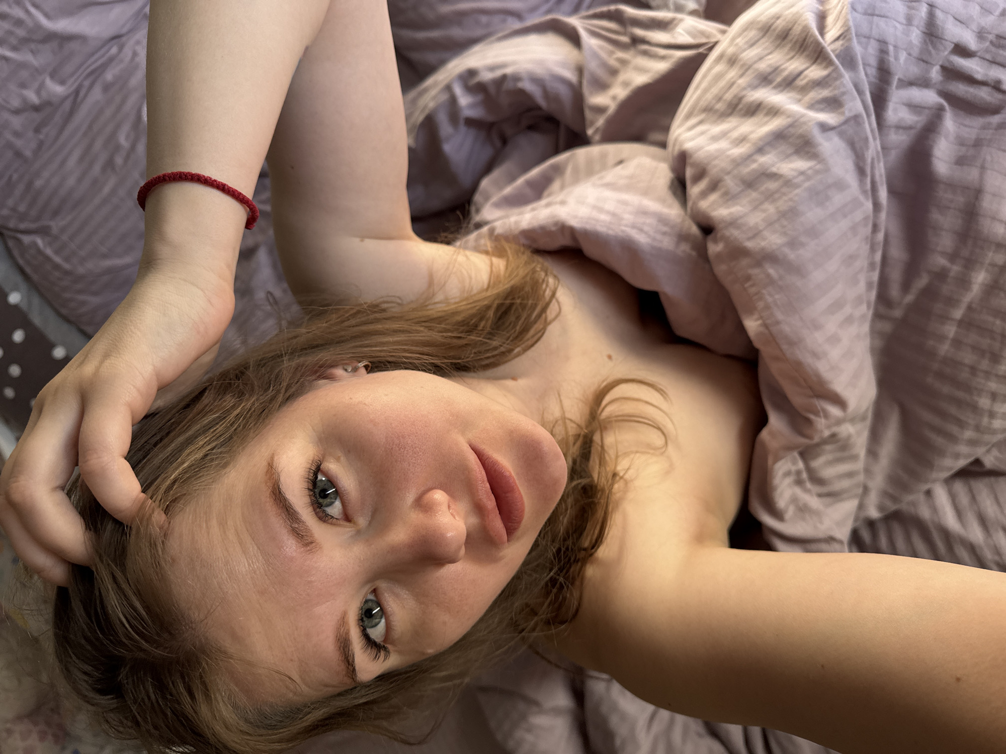 A fair-skinned woman with light brown hair lies in a bed covered with light purple striped sheets and a similar colored duvet. She is looking at the camera with a slight smile. A red woven bracelet is visible on her left wrist. Her left hand is touching her hair. The location is unspecified but may be in an indoor setting like a home or hotel, not a public space in Gornyi. No information is available to determine if this photo was actually taken in Gornyi (Dvurechenskoe S/P), Russia.