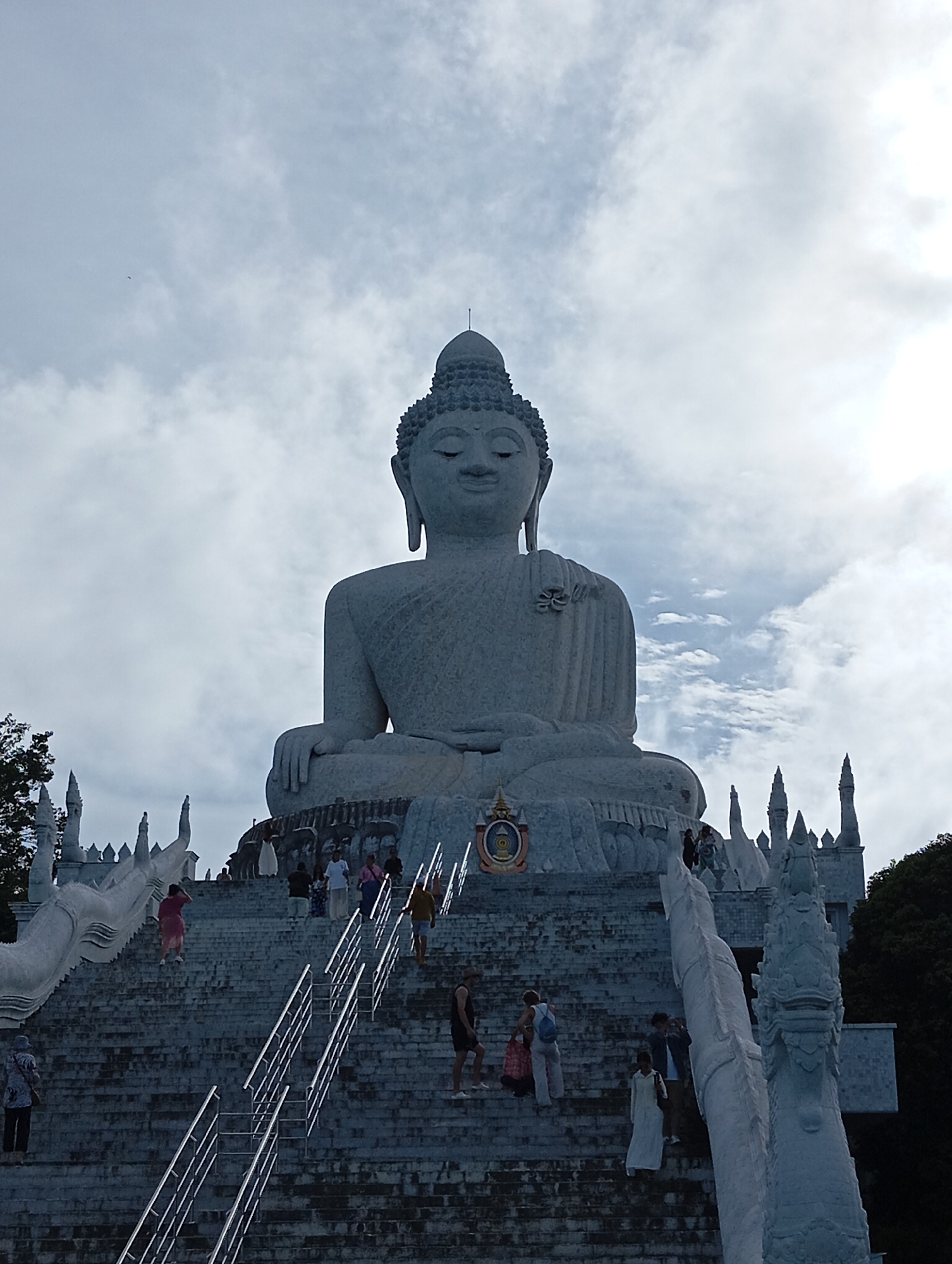 The image shows the Big Buddha statue in Karon, Thailand, under a cloudy sky. The large, white marble statue of Buddha is seated in a meditation pose, with his face looking serenely forward. The statue is perched atop a hill, with a grand staircase leading up to it. Numerous people are seen ascending and descending the stairs, dressed in casual clothing. The staircase is flanked by ornate balustrades shaped like mythical serpents. In the background, lush green trees are visible, contrasting with the white of the statue and stairs. The overall scene evokes a sense of religious pilgrimage and cultural tourism.