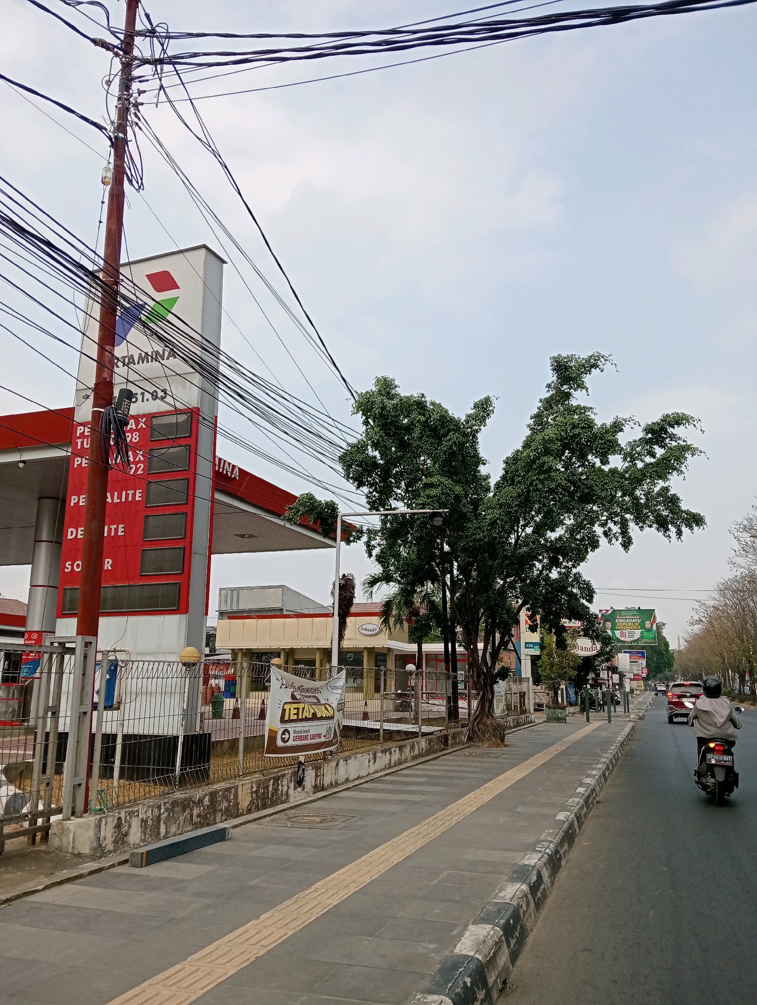 The image shows a street scene in Samarinda, Indonesia, featuring a Pertamina gas station on the left side. A tall pole with numerous tangled electrical wires is prominent in the foreground, extending across the top of the frame. The Pertamina sign is visible, displaying fuel prices on a red vertical panel with digital readouts. A sidewalk runs alongside the gas station, separated from the road by a curb. A person on a motorcycle is riding away from the camera on the right side of the road. The road itself is asphalt, with white and black markings on the curb. Trees line the right side of the street, and in the background, several buildings and billboards are visible. The sky is overcast.