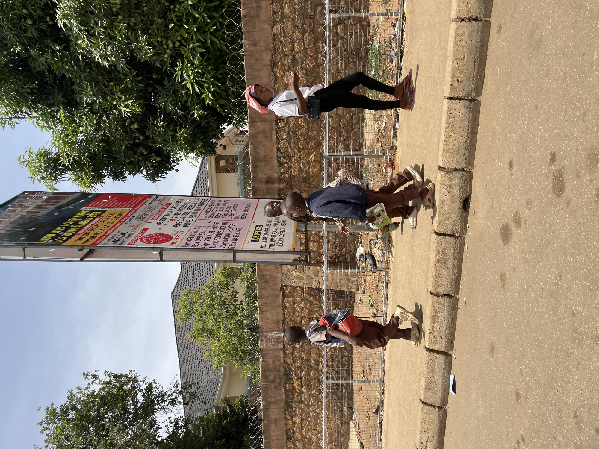 The image shows a street scene in Bukuru, Nigeria. Three people are visible along the sidewalk. A woman in a white shirt, black leggings, and a headscarf is standing and looking at her phone. Two boys in school uniforms are further down the sidewalk; one is walking, and the other is standing. A large sign advertising "Lockup Shop" and "Warehouse" is on the left side of the image. The background includes a stone wall with barbed wire, trees, and buildings. There is visible litter and trash along the fence in the background.