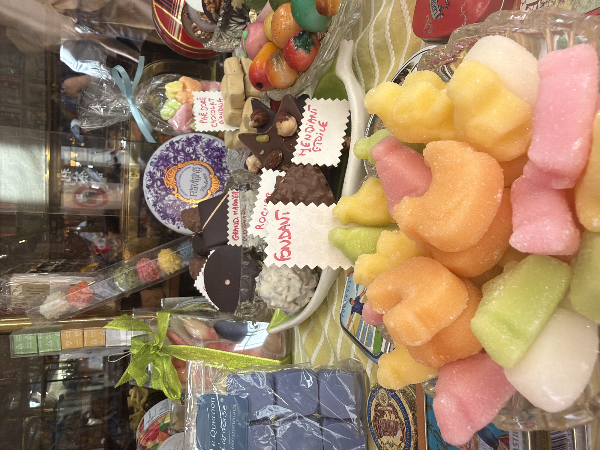 The image shows a display case filled with various French sweets and candies. Among the items visible are candied fruits, chocolate assortments, and sugar-coated candies in various shapes and colors. Some of the candies are labeled with French names such as "Pave Dore Chocolat Gianduja," "Le Quernon L'ardoise Chocolat et Nougatine," "Grand Harmer," "Roche Fondant," and "Mendiant Etoile." There is also a tin of "Les Anis de Flavigny Violette" candies. The display suggests a specialty candy shop in Paris, France.