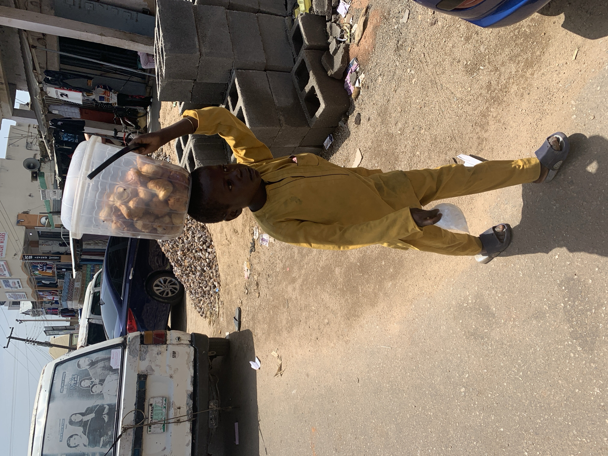 In Jos, Nigeria, a young boy, likely a vendor, walks down a sunlit street carrying a large clear plastic bucket filled with food items on his head. He is dressed in a yellow traditional African outfit and wears dark sandals. In his right hand, he holds a white plastic bag. The street is paved with asphalt and is bordered by a dirt shoulder.
To the left of the boy, a white van with a license plate reading "PLATEAU 91VE" is parked. The van's rear is weathered, and a poster advertising "NNAJI MOVIES" is visible on its back. Behind the van, a dark blue car is parked.
To the right of the boy, a stack of gray concrete blocks forms a makeshift construction or storage area. Scattered around the blocks are various debris, including pieces of plastic and paper. In the background, buildings line the street, displaying shops with hanging clothing and signs, suggesting a commercial district. A utility pole with wires stands tall against the bright sky. The overall scene depicts a bustling urban environment in Jos, with signs of commerce and daily life.