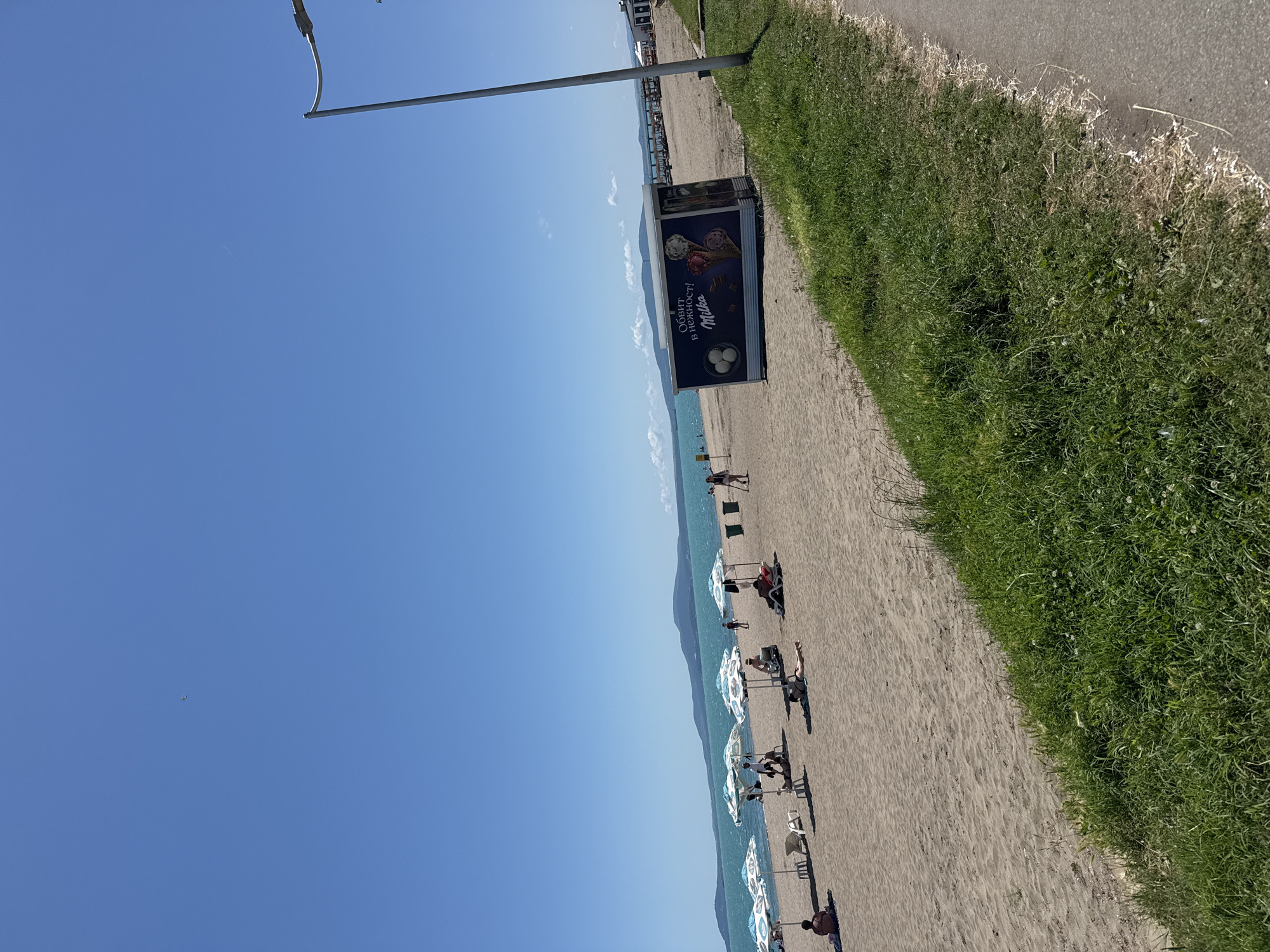 The image depicts a sunny day at a sandy beach, likely in Burgas, Bulgaria, given the presence of a Milka ice cream kiosk. The scene is framed by a clear blue sky, with a faint contrail from an airplane visible. The beach is lined with several blue and white patterned umbrellas, offering shade to beachgoers. Some people are seen relaxing on lounge chairs or directly on the sand.
In the foreground, lush green grass lines the edge of a paved pathway, suggesting a promenade or sidewalk adjacent to the beach. Further down the beach, a small, dark blue kiosk with advertisements for Milka ice cream, featuring cones and spheres, is positioned. The sea is calm and turquoise, meeting a distant coastline with rolling hills. A tall lamppost stands on the right side of the image. Two trash cans are visible near the kiosk. The overall atmosphere is one of a typical summer day at the seaside.