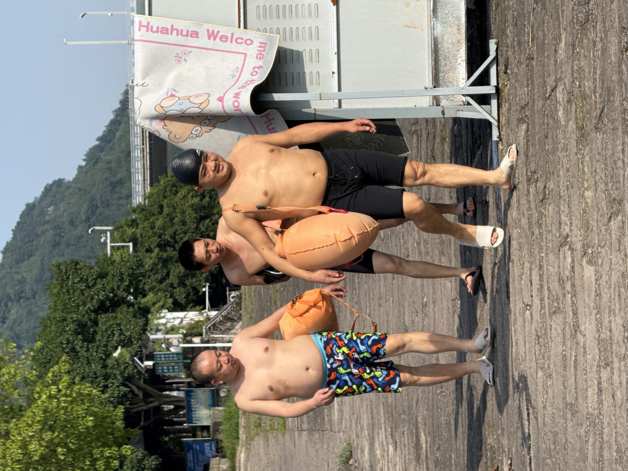 The image shows three East Asian men outdoors in what appears to be Jiangjin, China, dressed in swimwear and holding orange inflatable buoys, suggesting they are engaging in or preparing for open-water swimming. They are standing on a light-colored, textured paved surface, likely a riverside or lakeside path.
The man in the foreground on the left wears colorful patterned swim shorts and holds an orange buoy. The central man wears black swim shorts and also carries an orange buoy. The man on the right, also in black swim shorts, wears a black swimming cap and holds an orange buoy, smiling broadly. All three men are shirtless and wearing sandals.
The background features lush green mountains and trees under a clear blue sky, indicating a bright and sunny day. Strong shadows are cast on the ground, suggesting it is daytime, possibly mid-day. A metal structure is visible behind the men, from which hangs a white banner with pink text. Partially visible text on the banner reads "Huahua Welco" and "me to the Wo," along with a cartoon character design. The scene conveys a recreational outdoor activity in a scenic, natural environment.
