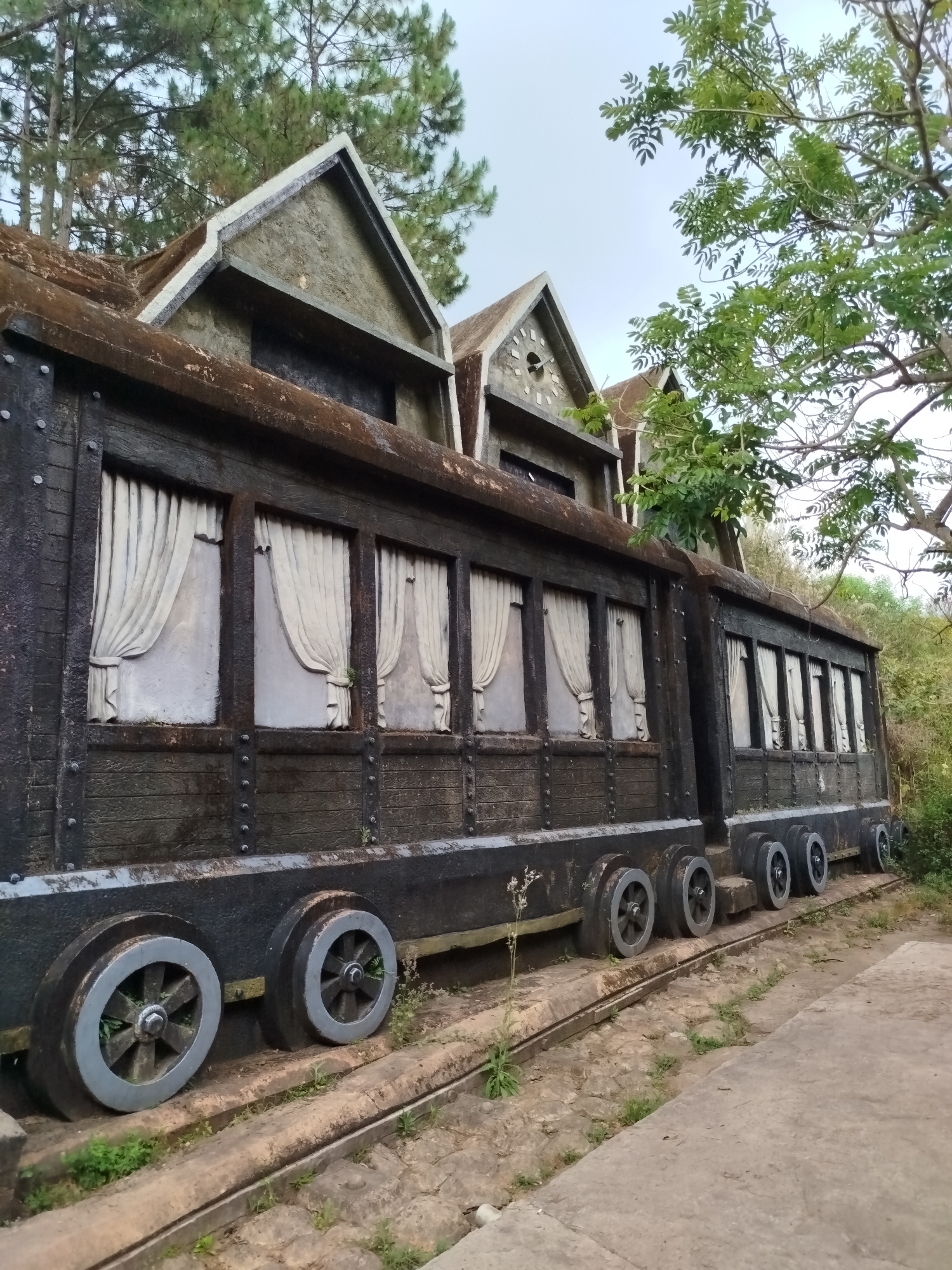 The image shows a static, simulated train car structure, constructed of a dark, textured material, possibly concrete, mimicking a vintage train car. The structure is positioned on a mock rail track, with prominent wheels fabricated to resemble those of a train. The car features a series of framed window spaces with molded white curtains, suggesting a passenger compartment. Above the car are architectural additions that resemble building facades, topped with peaked roofs, adding a whimsical element. The structure is situated outdoors, surrounded by greenery, including trees and sparse vegetation. The ground is a mix of concrete and natural earth. The sky is overcast, indicating a diffuse lighting condition. The overall scene is located in Da Lat, Vietnam, based on common knowledge of similar tourist attractions in the area.
