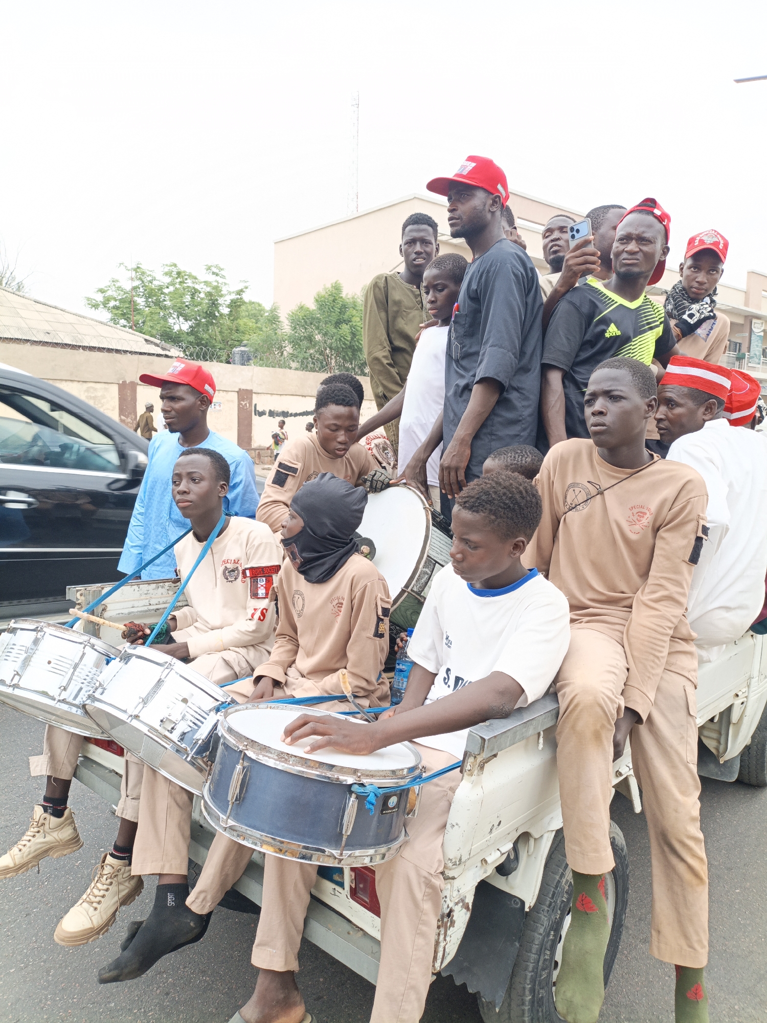 A group of youths are crowded onto the back of a white truck on a street in Tudun Wada, Nigeria. Several are wearing red hats. Three young men sit in front, playing snare drums that are suspended by blue straps. They are wearing uniforms that include patches and emblems. Others are standing behind them and looking around; some are holding cell phones. A dark-colored sedan is visible in the foreground to the left. Buildings and trees are visible in the background.