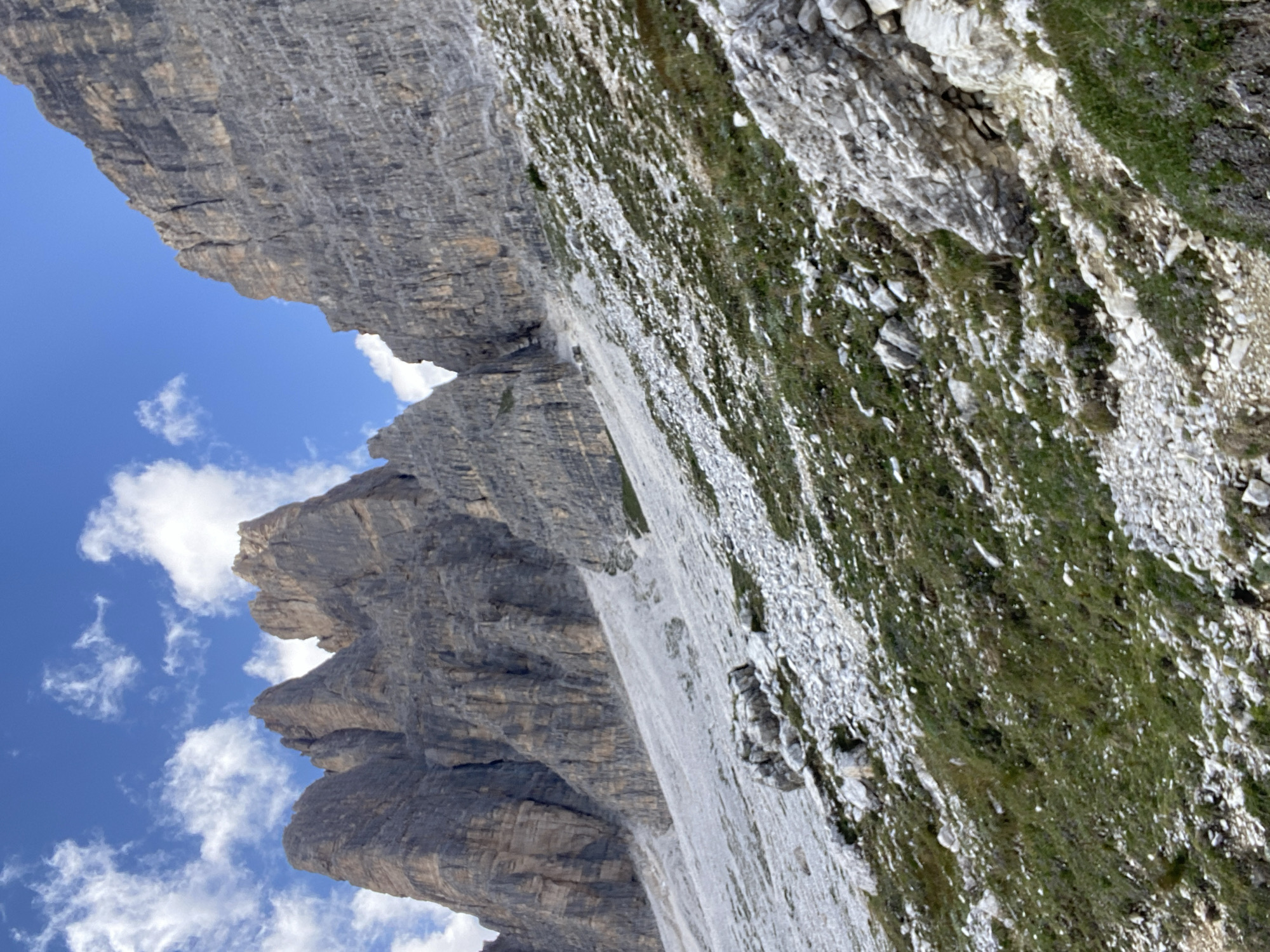 The image depicts a rugged mountainous landscape under a bright blue sky dotted with white clouds. The foreground features a steep, rocky slope covered with sparse green vegetation and scattered white stones. The midground showcases a large scree slope, a steep incline composed of loose rocks and gravel, which transitions into the imposing mountain formations in the background. These mountains are characterized by their sheer, grey rock faces, exhibiting deep crevices and layered textures. The overall scene conveys a sense of natural grandeur and wilderness, typical of alpine environments. While the image is of a mountainous region, no specific landmarks are visible to pinpoint the exact location within Auronzo di Cadore, Italy, though it is consistent with the area's known topography. No people, animals, or human-made structures are present in the image.